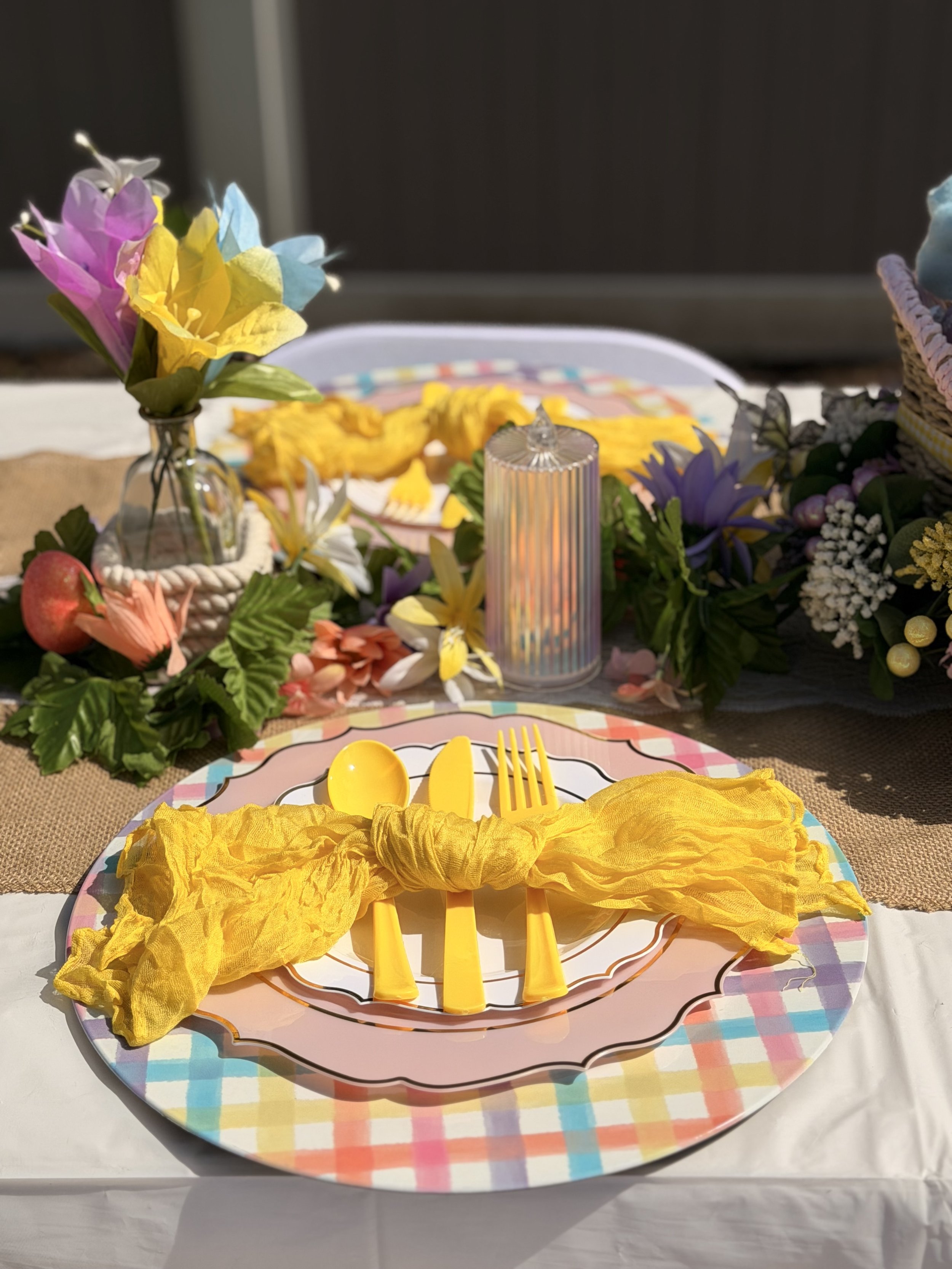 Colorful table setting with a pink and checkered plate, yellow cutlery tied with a yellow cloth, surrounded by flowers and decorations.