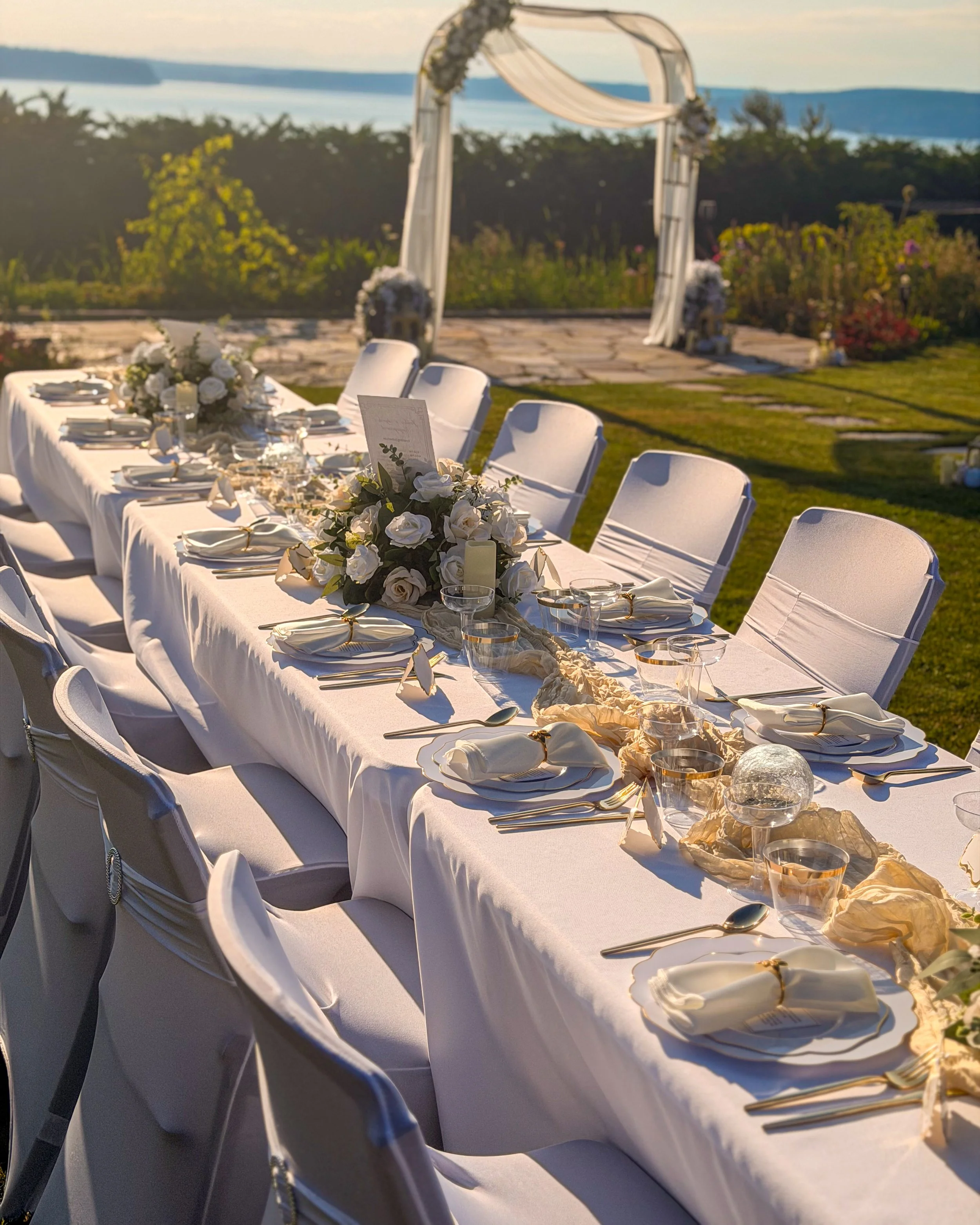 Wedding reception table outdoors with white chairs, floral centerpieces, plates, and cutlery, overlooking a scenic body of water and distant hills at sunset.