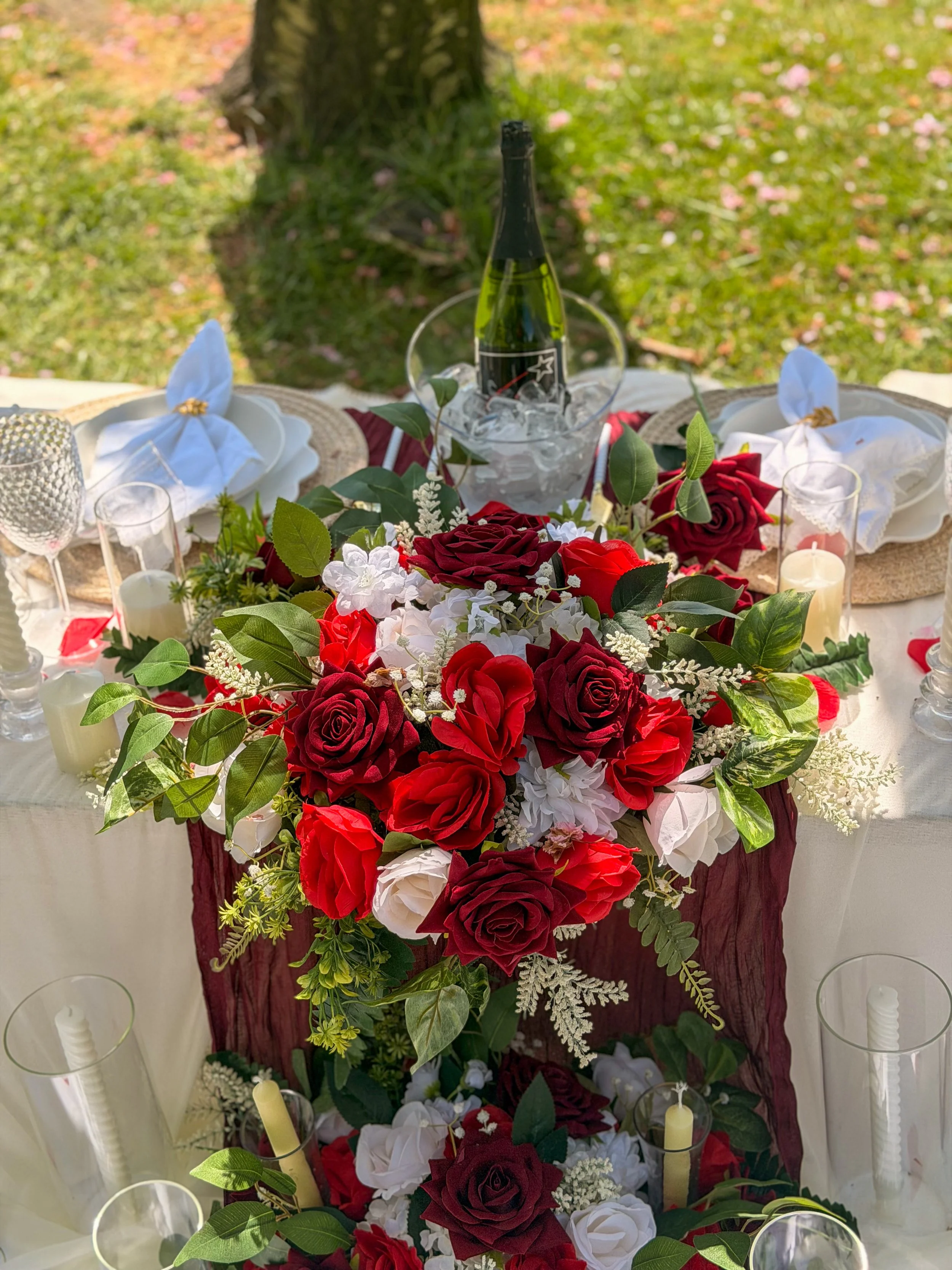 A decorated table for a celebration with a floral centerpiece, a bottle of champagne on ice, candles, and neatly folded napkins in a scenic outdoor setting.