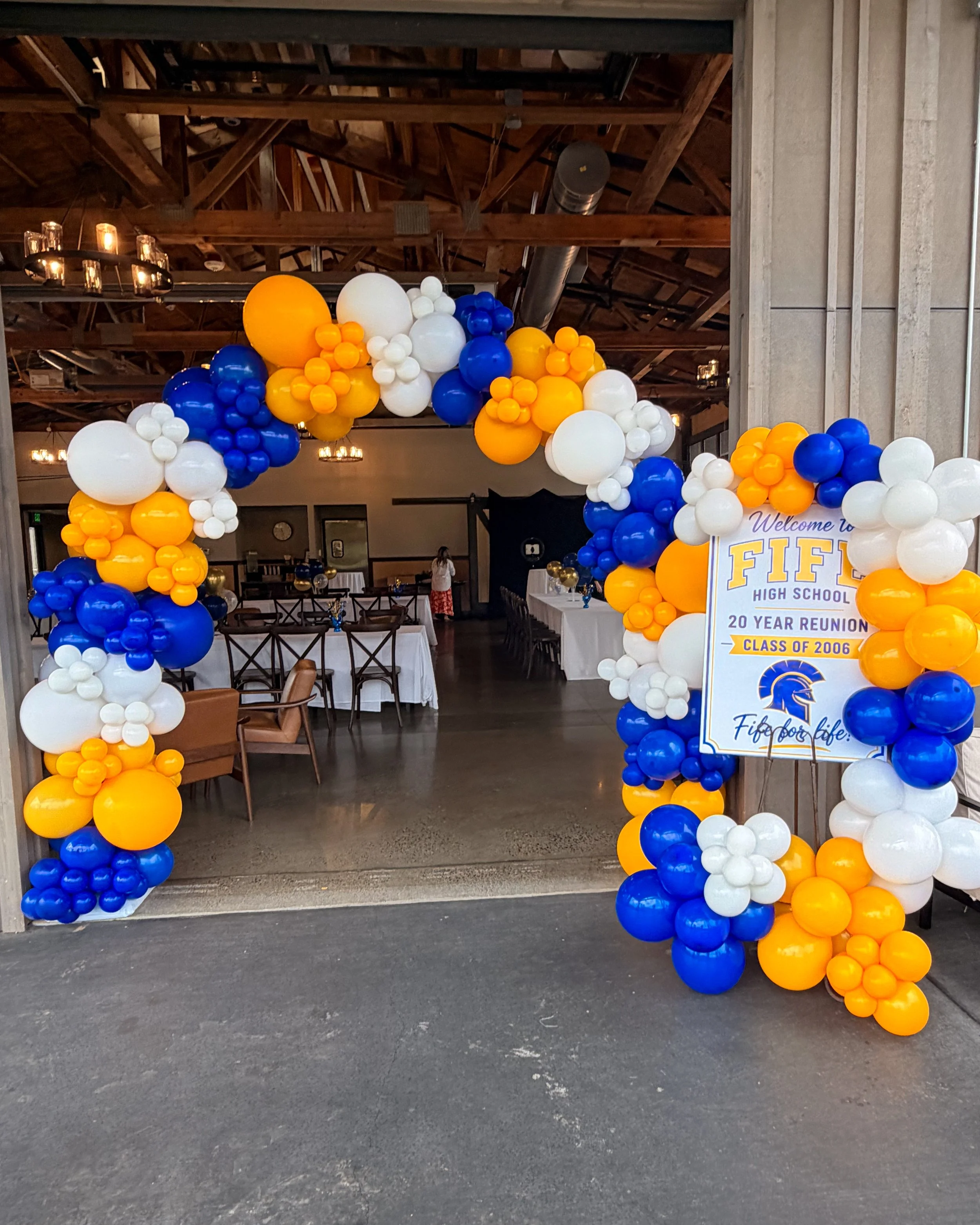 Decorative balloon arch in blue, yellow, white, and orange colors for a high school reunion at FIFE High School, with tables and chairs set up inside the venue and a person walking in the background.