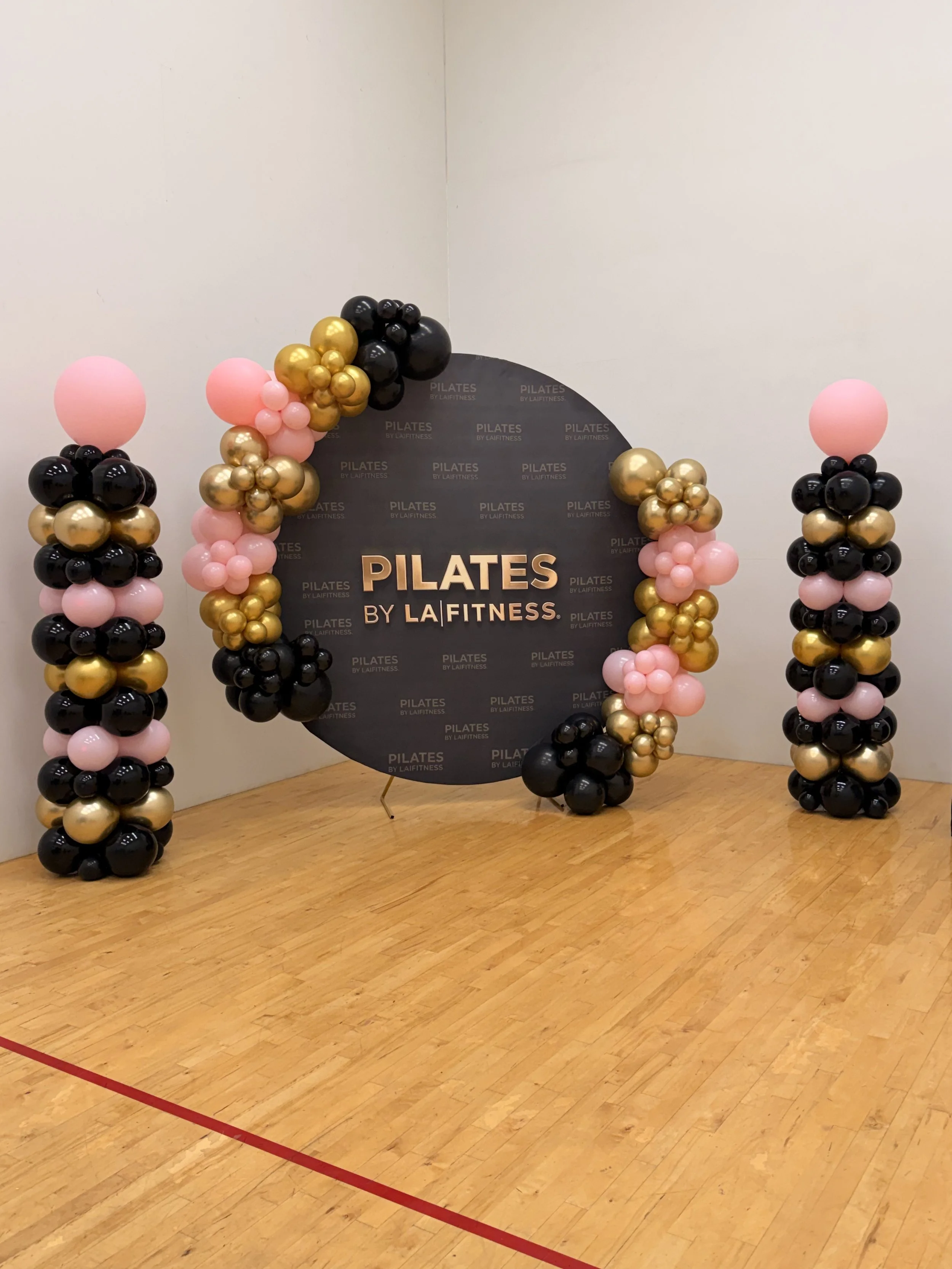 Balloon arches with black, pink, and gold balloons surrounding a Pilates promotional backdrop in a gym or fitness studio.