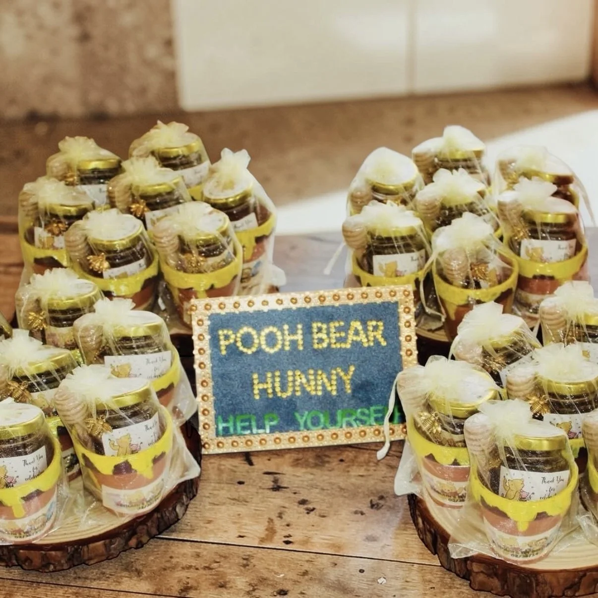 Small jars with Pooh Bear labels, decorated with yellow ribbons and white tulle, arranged on wooden slabs, with a handwritten sign reading "Pooh Bear Hunny Help Yourself".