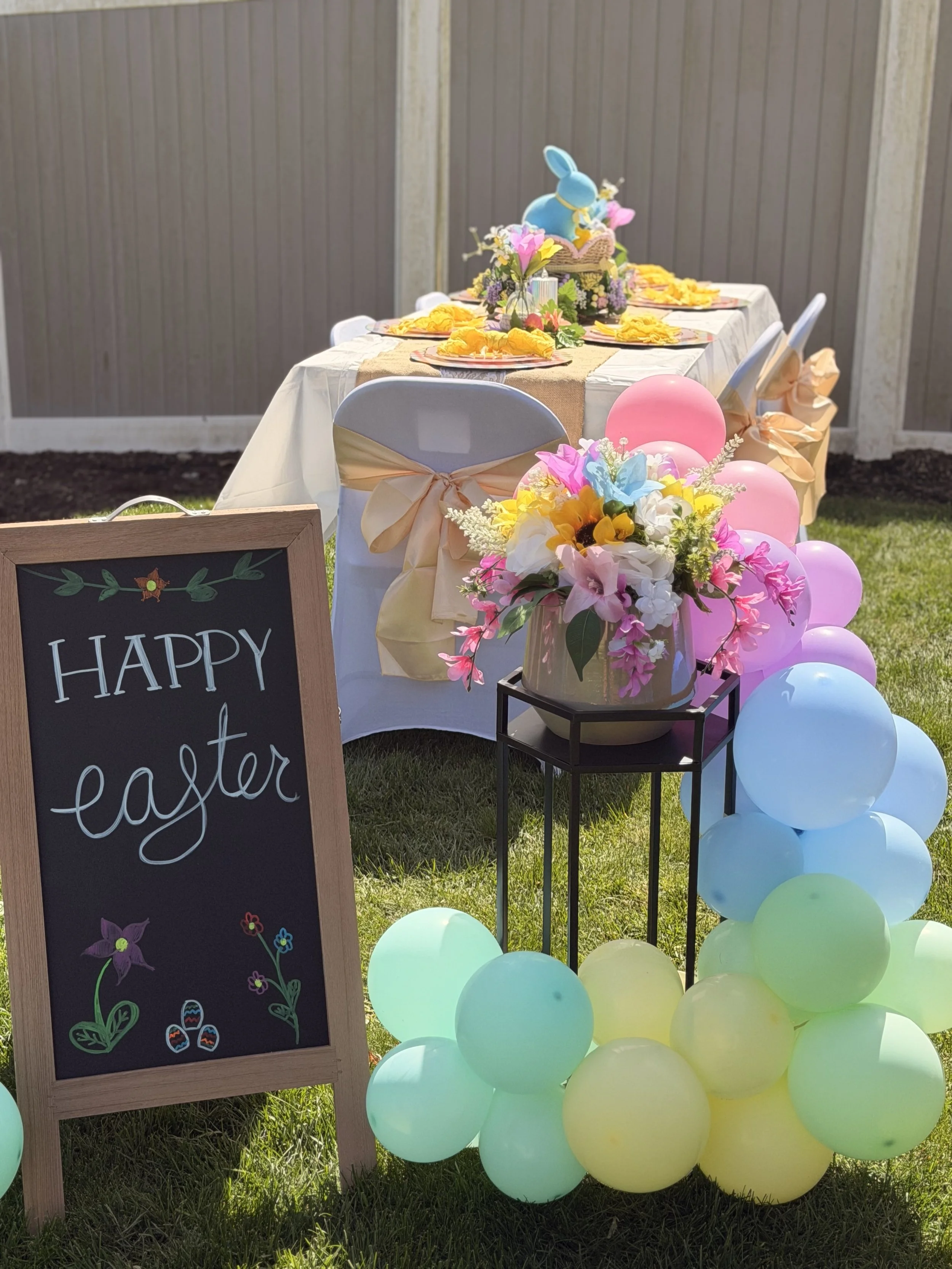 Easter outdoor celebration setup with a decorated table, pastel balloons, a floral arrangement, and a chalkboard sign that reads 'Happy Easter'