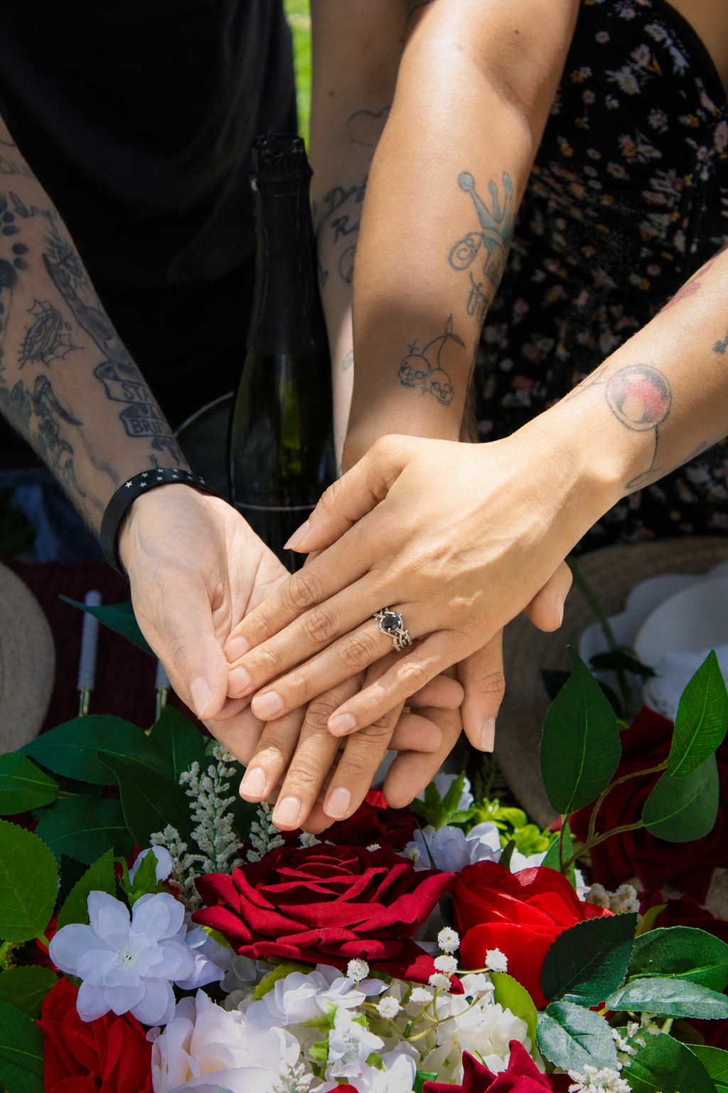 Three people with tattooed arms join hands over a bouquet of red and white flowers, with a champagne bottle in the background.