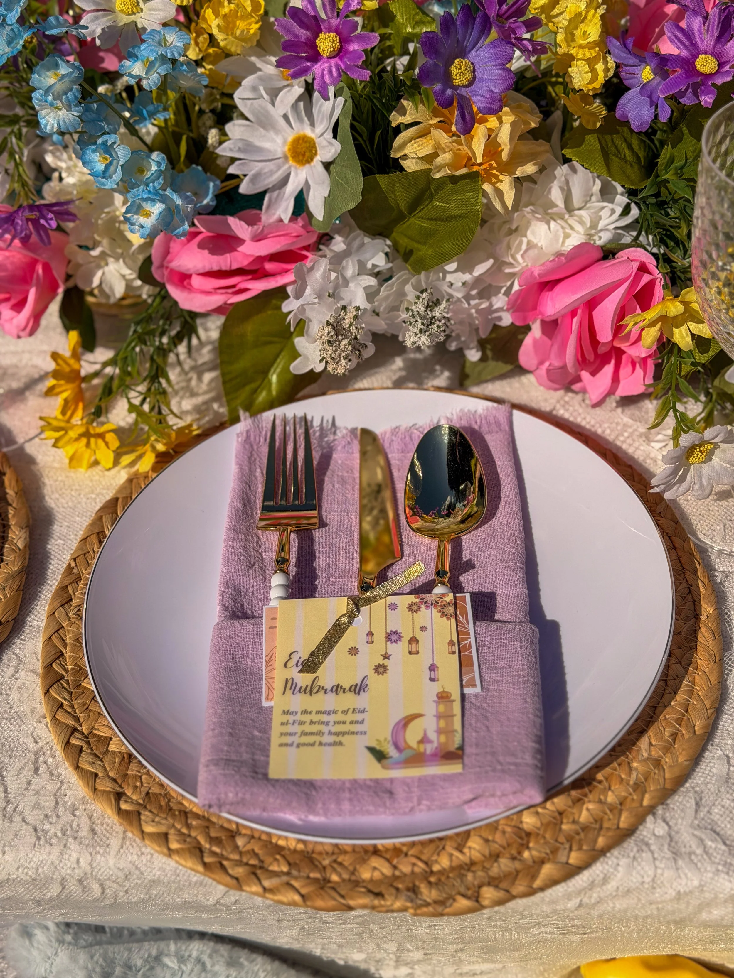 A table setting with a white plate on a pink napkin, gold fork and spoon, a decorative card with Eid Mubarak message, and a floral arrangement with white daisies, pink roses, purple and blue flowers, and yellow accents.