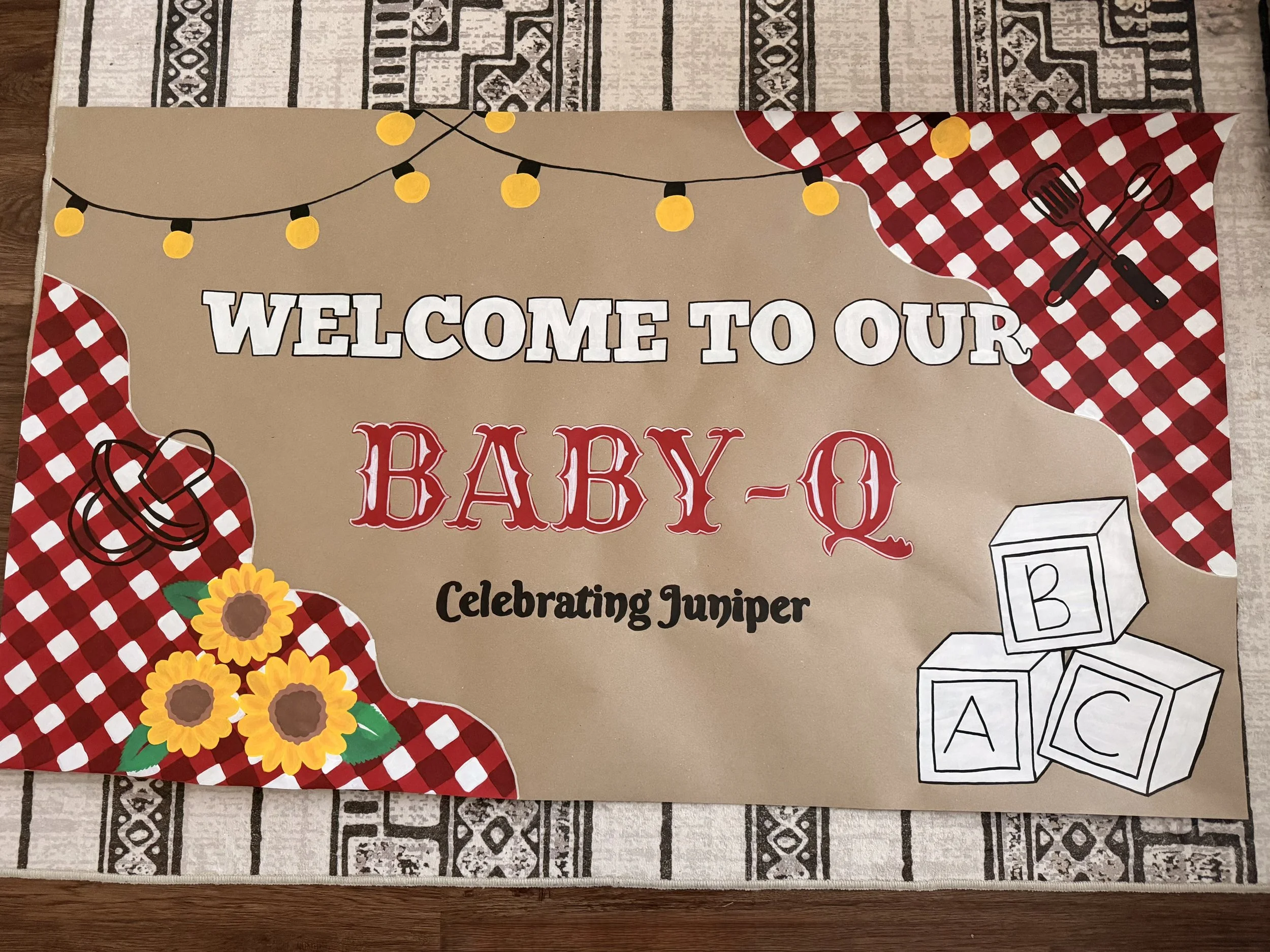 A welcoming sign for a baby shower with a picnic theme, featuring a string of yellow lights, sunflower decorations, red and white checkered accents, and illustrations of children's blocks and cooking utensils.