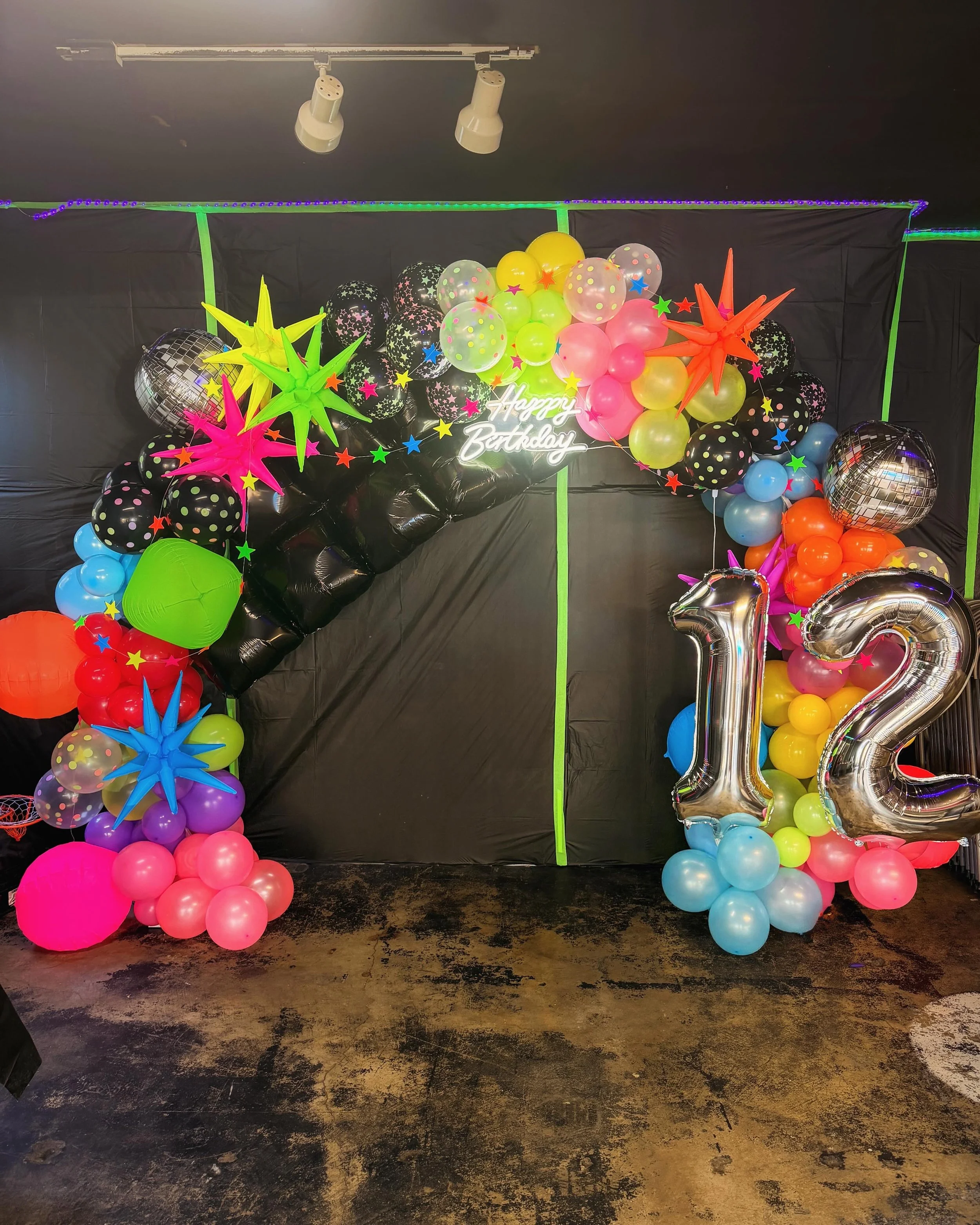 Colorful birthday party decoration with balloons, star-shaped ornaments, a neon 'Happy Birthday' sign, and large silver balloons forming the number '12', all set against a black backdrop.