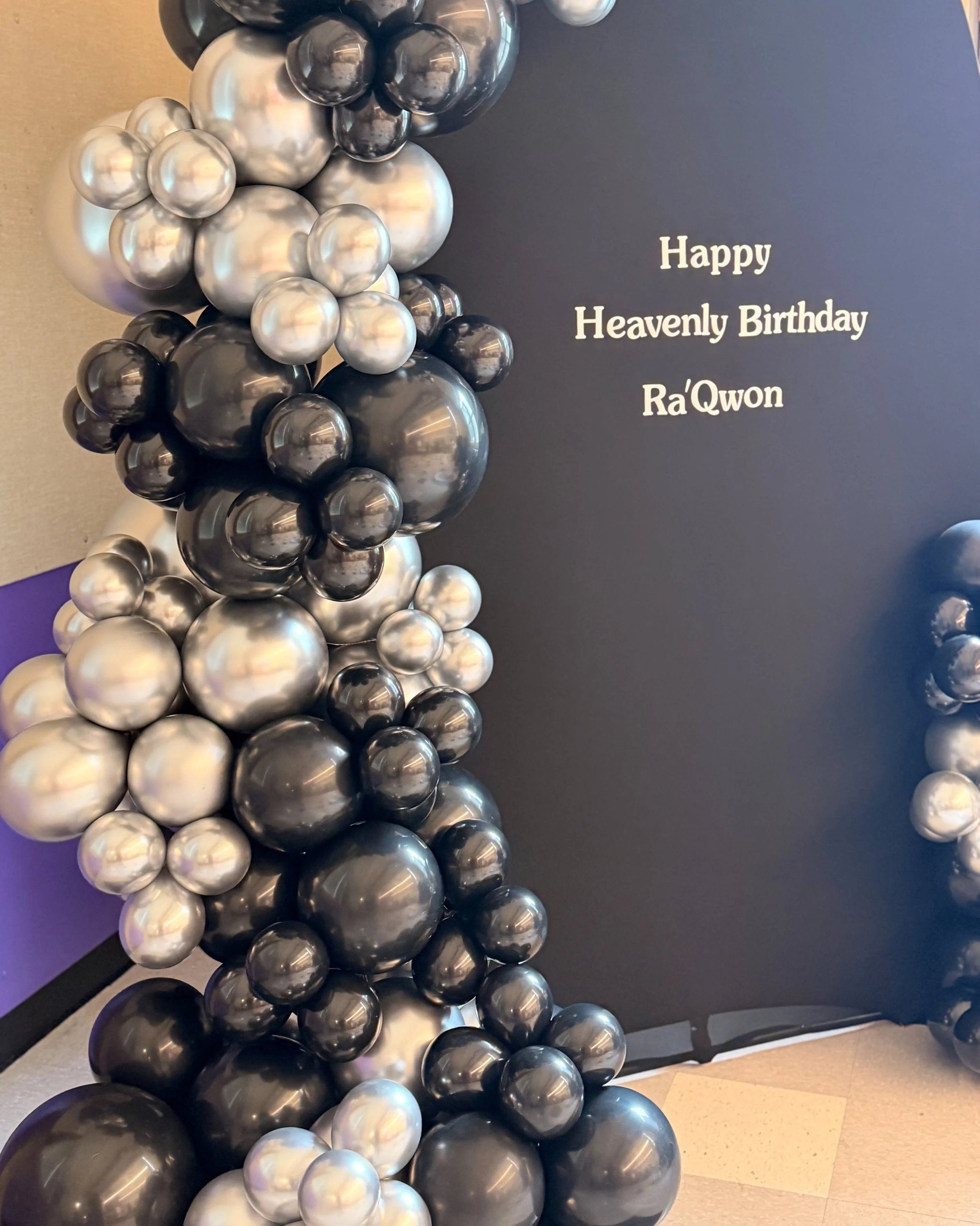 A balloon sculpture made of black, gray, and white balloons arranged in a pattern on a dark wall. To the right of the sculpture, a wall reads 'Happy Heavenly Birthday Ra'Qwon.'
