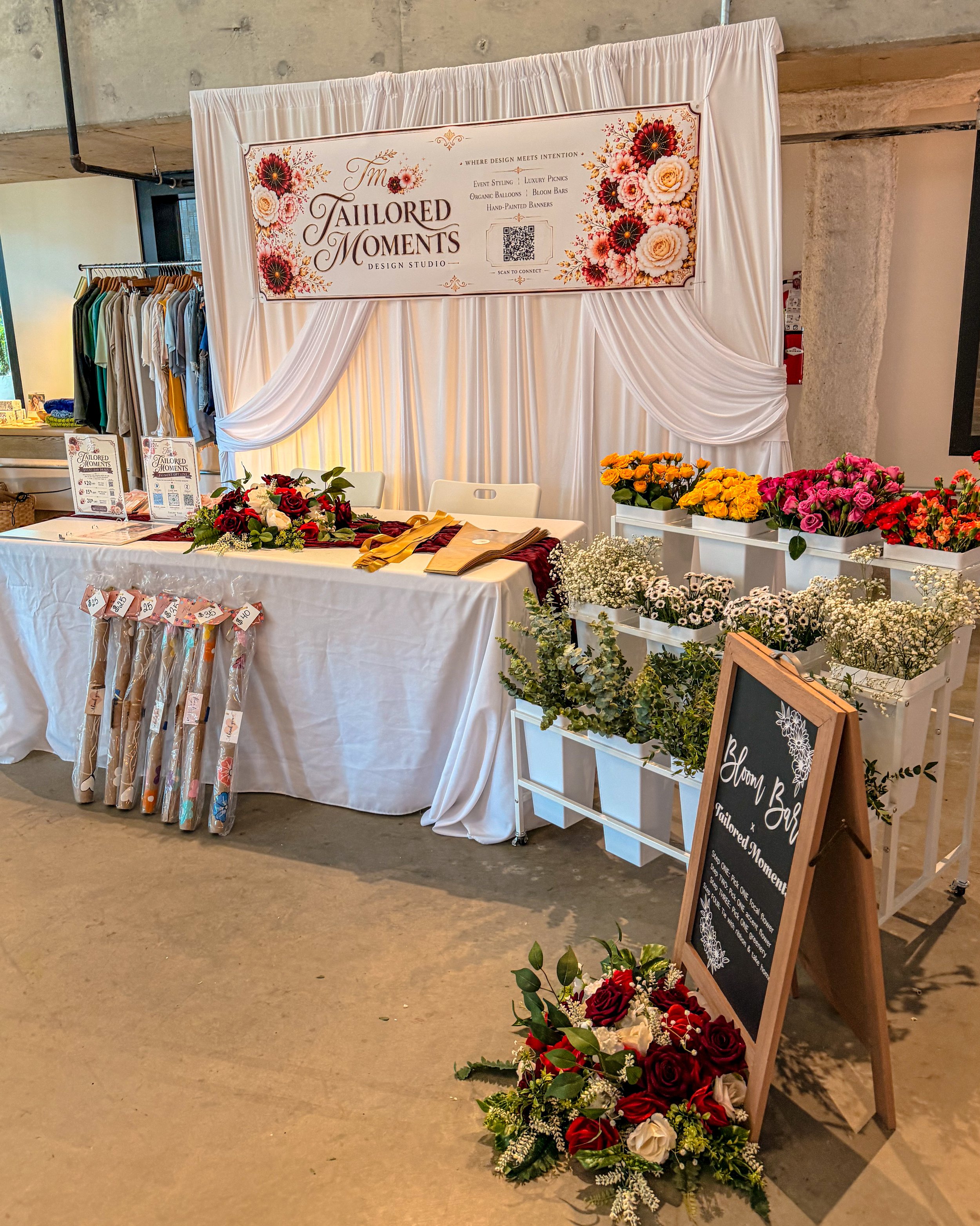 A wedding or event decor booth titled 'The Tailored Moments Design Studio' featuring a white draped backdrop with a sign decorated with red, pink, and white flowers. The booth displays floral arrangements in baskets and a chalkboard sign, with flower