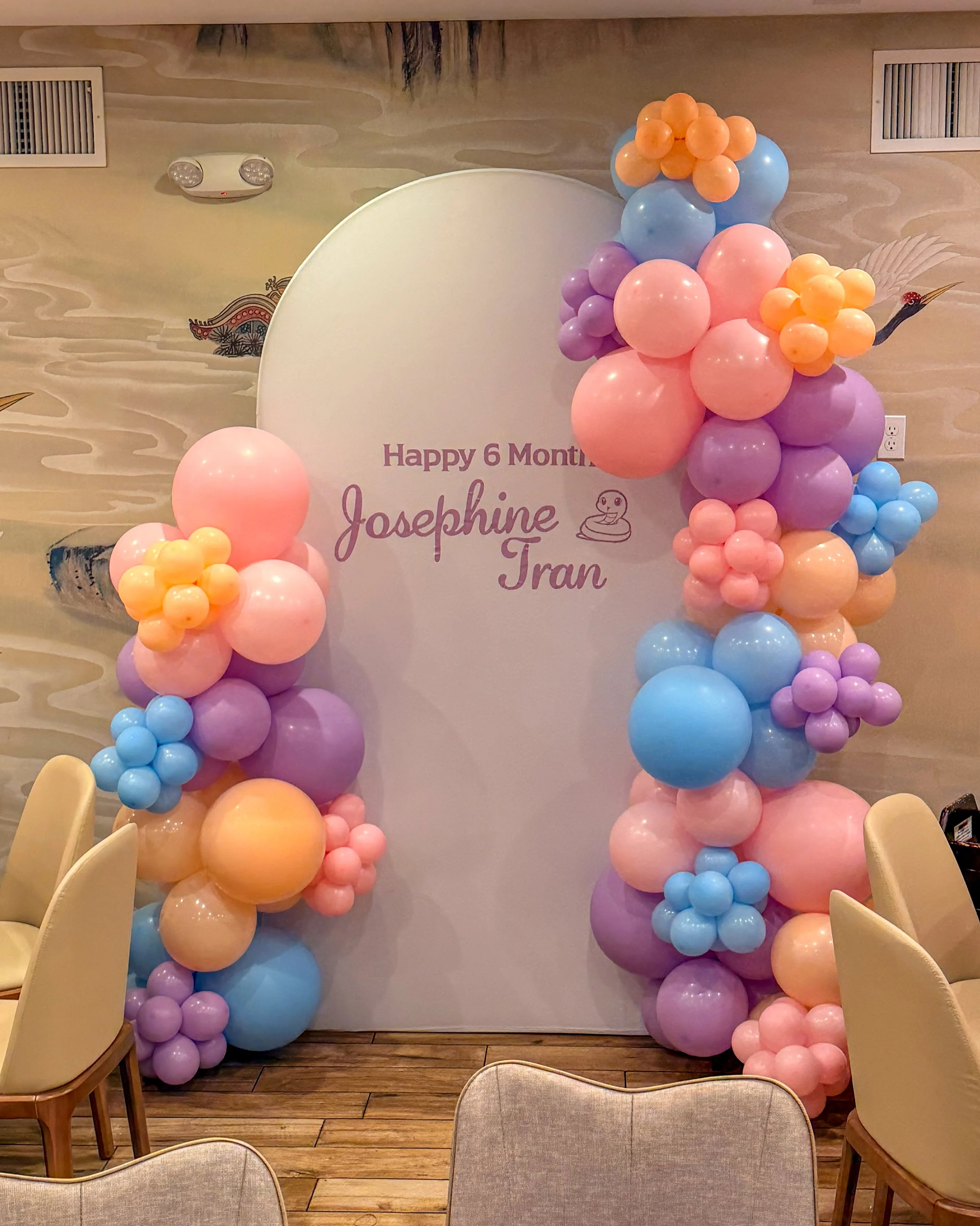 Decorative balloon arch in pastel colors with pink, purple, blue, and peach balloons, set against a wall with a scenic mural, part of a celebration setup for a child's sixth month milestone.