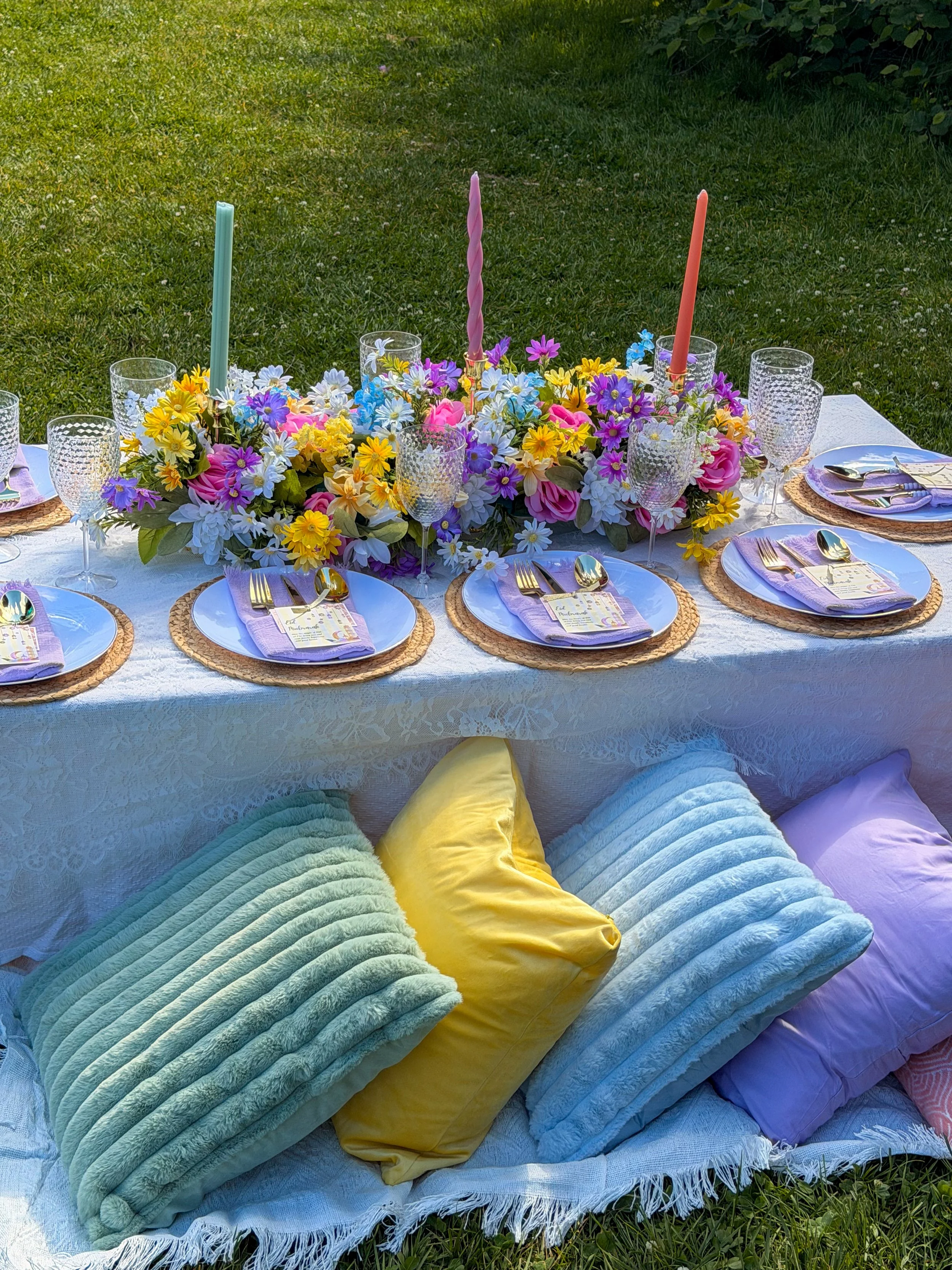 A table set outdoors with colorful flowers as a centerpiece and place settings including plates, glasses, utensils, and cloth napkins. Cushions are arranged on the ground around the table on a grassy area.