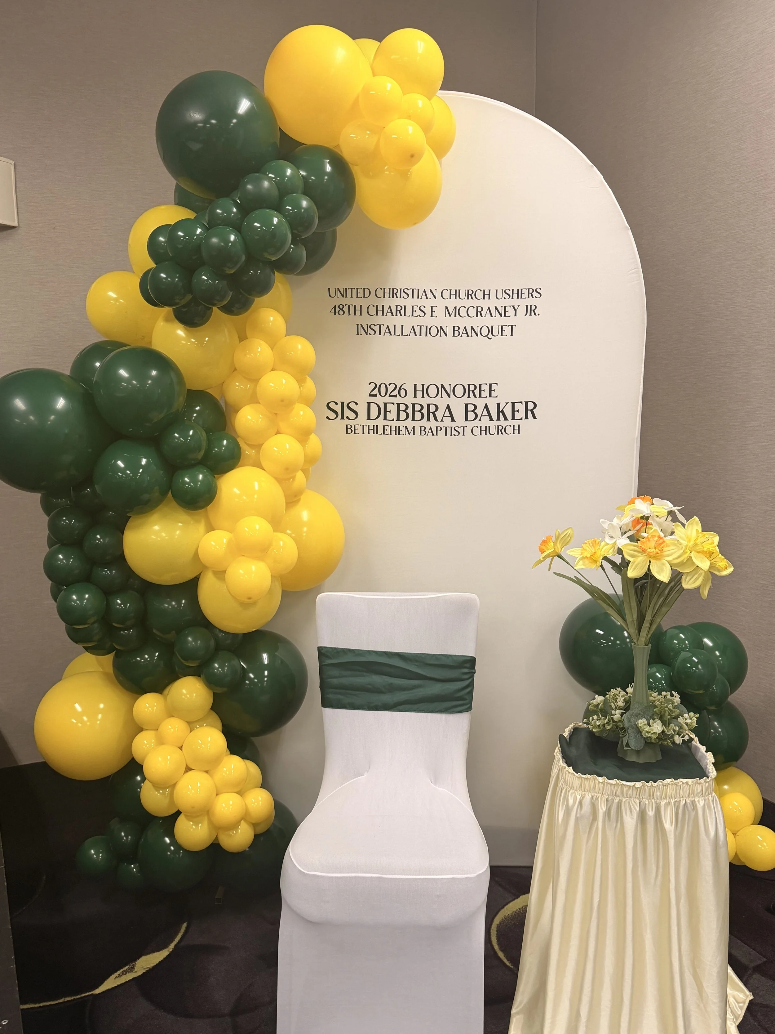 Decorative setup with yellow and green balloons, a white chair, and a floral arrangement with yellow and white flowers, for an event honoring Sis Debbra Baker at Bethlehem Baptist Church.