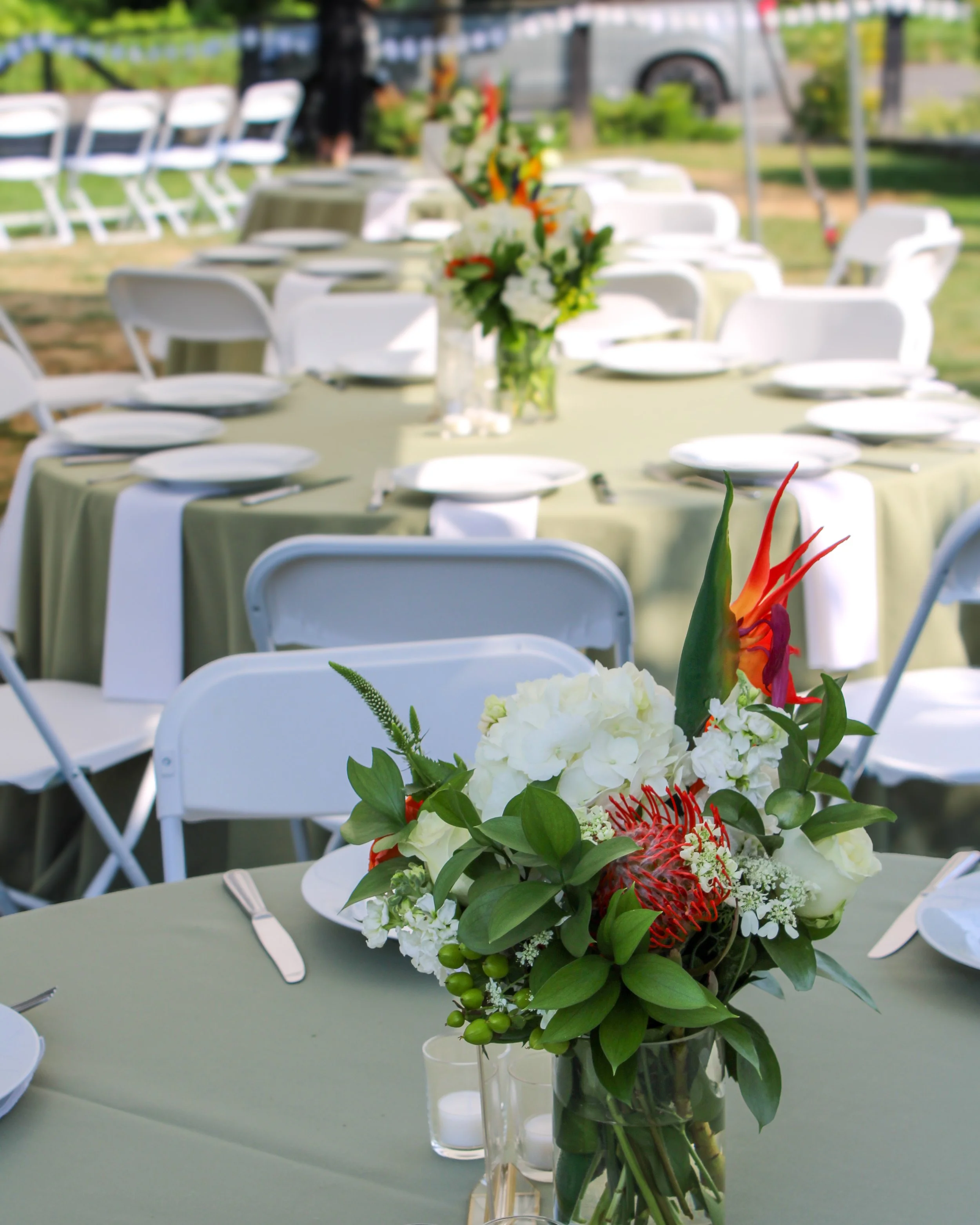 A floral centerpiece with white, orange, and green flowers on a banquet table set for an outdoor event.