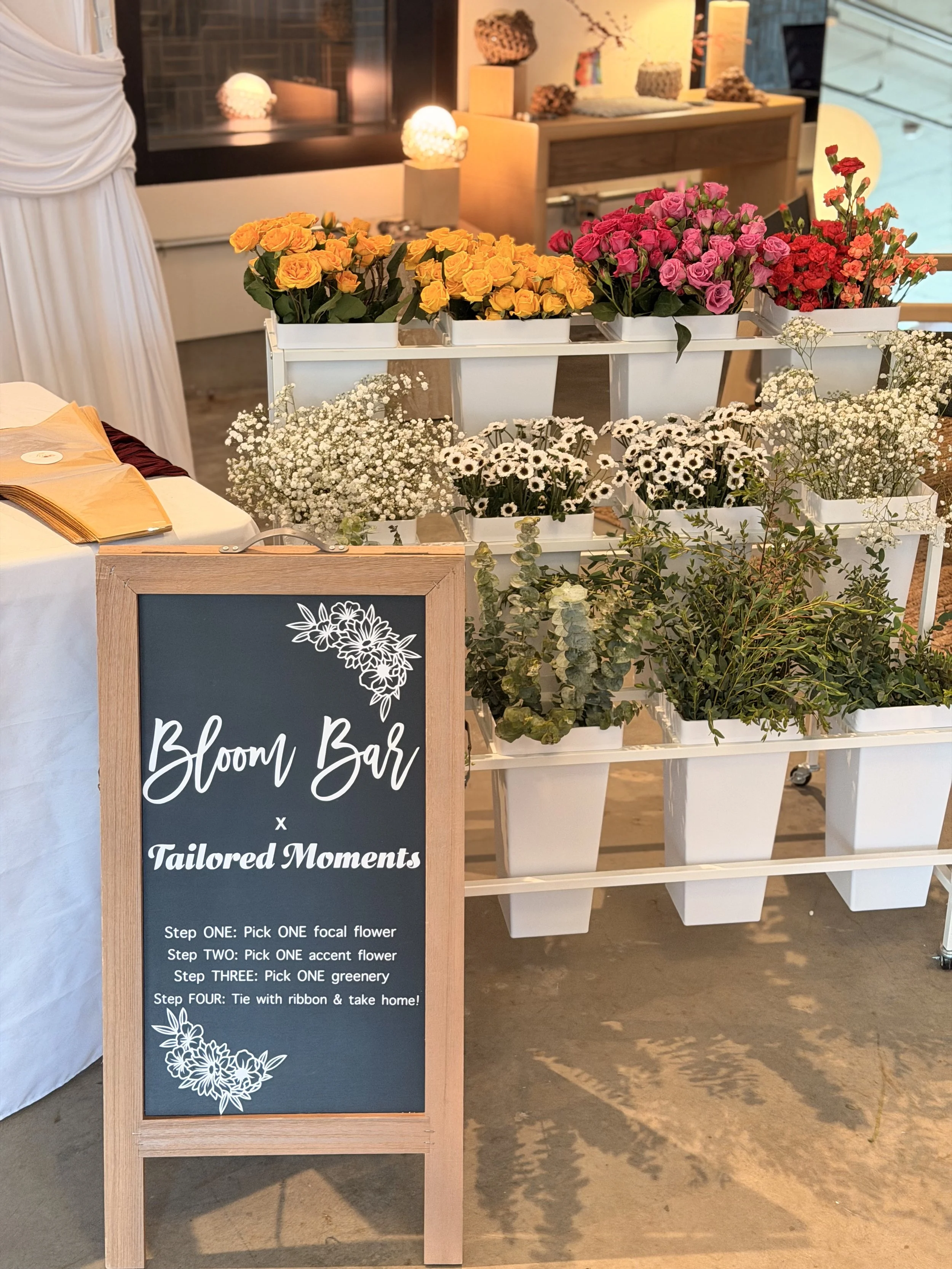 A chalkboard sign with the words 'Bloom Bar x Tailored Moments' and instructions for creating a floral arrangement next to a display of various colorful flowers in white pots, including yellow, pink, red, and white flowers, in a well-lit indoor space