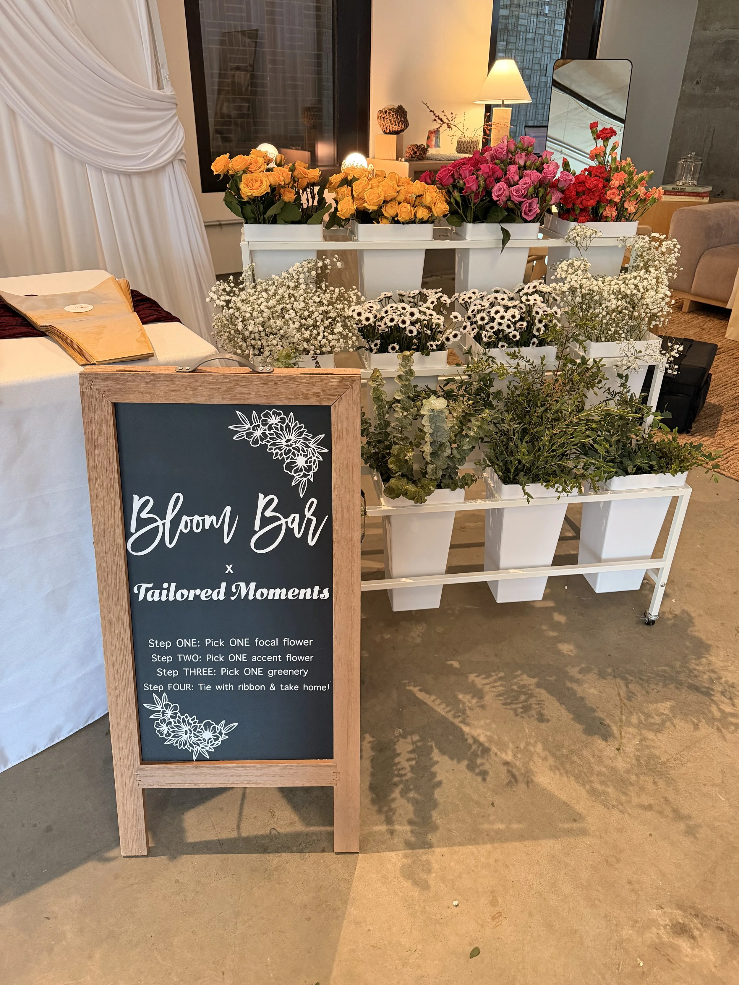 A blackboard sign with white floral decorations and the words 'Bloom Bar x Tailored Moments' written on it. Behind the sign, there are white shelves filled with various colorful flowers, including yellow roses, pink roses, red and pink carnations, white daisies, and baby's breath. There is also a table with a dark red cloth, a book or menu, and some decorations in the background, along with warm lighting and a mirror.