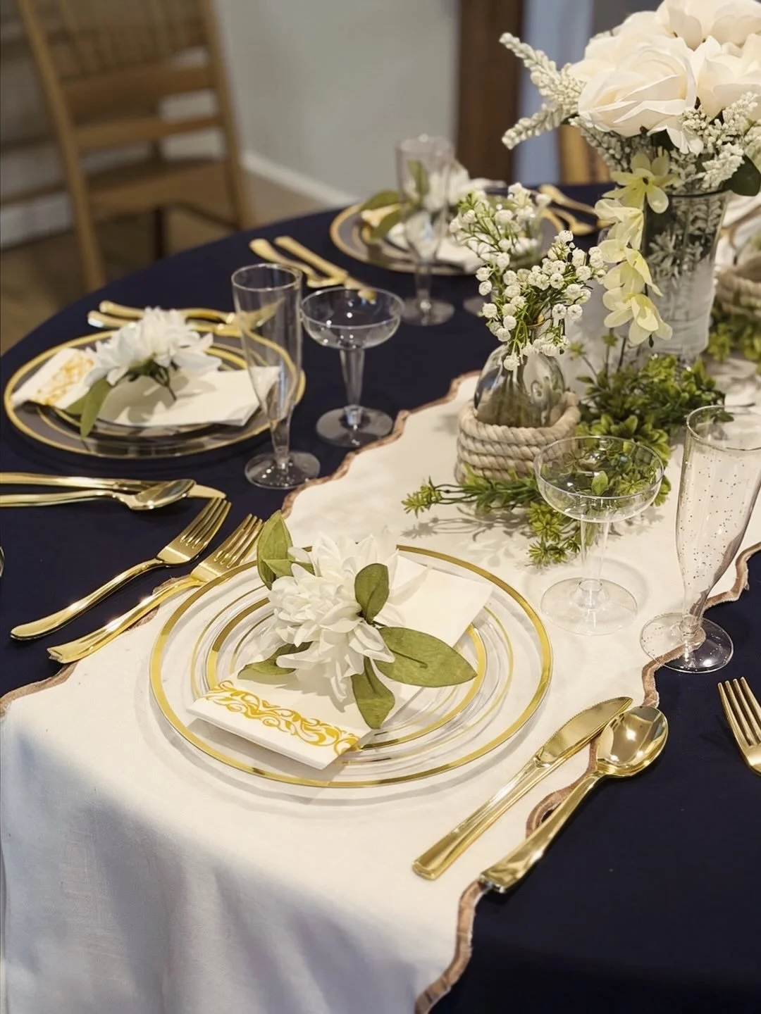 Elegant table setting with navy blue and white tablecloths, gold flatware, clear glassware, and floral centerpieces with white flowers and greenery.