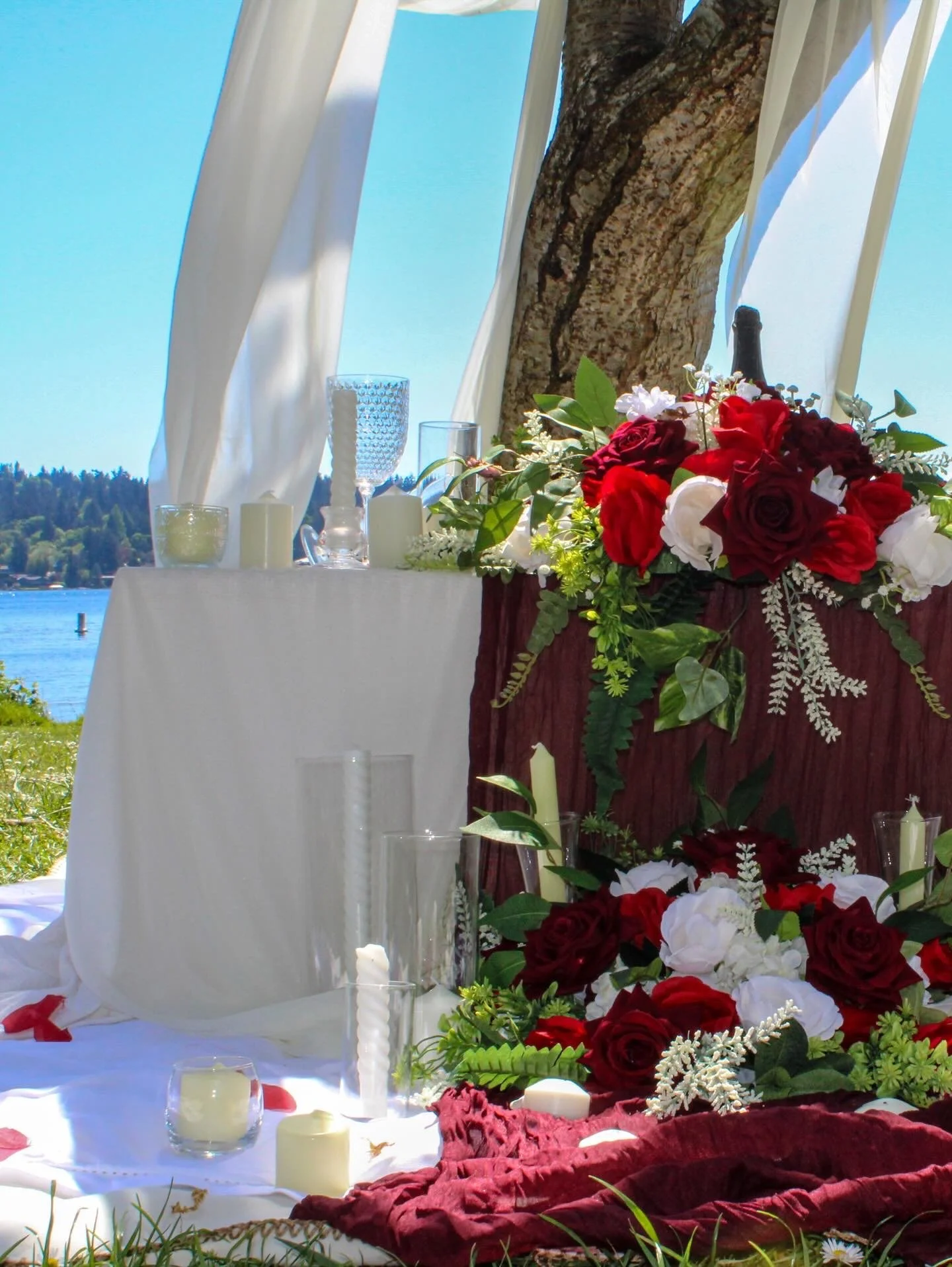 Decorative table setting outdoors with flowers, candles, glasses, and a bottle, near a large tree and a lake in the background.