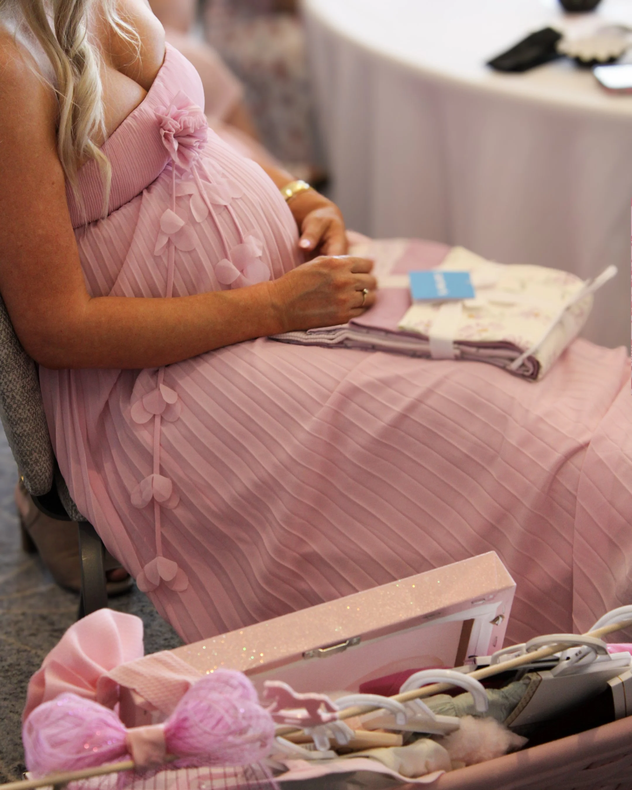 Close-up of a pregnant woman in a pink dress sitting at a table with pink yarn and other crocheting or knitting supplies.