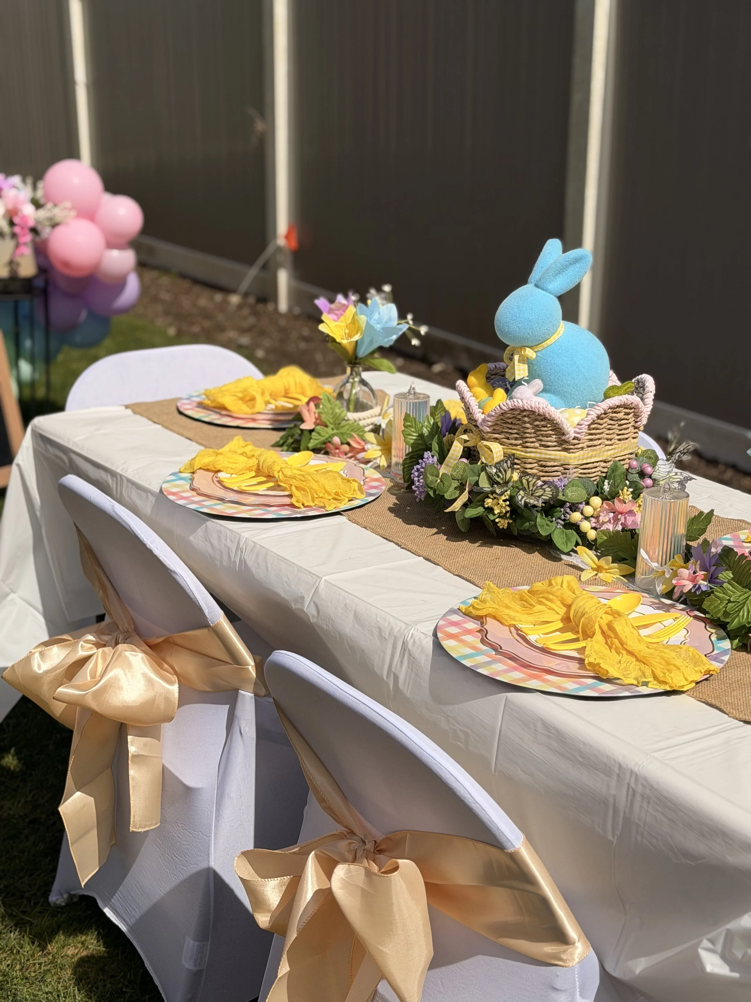 Easter-themed outdoor table setting with a stuffed blue bunny in a basket, decorated with flowers and yellow napkins, set for a celebration.