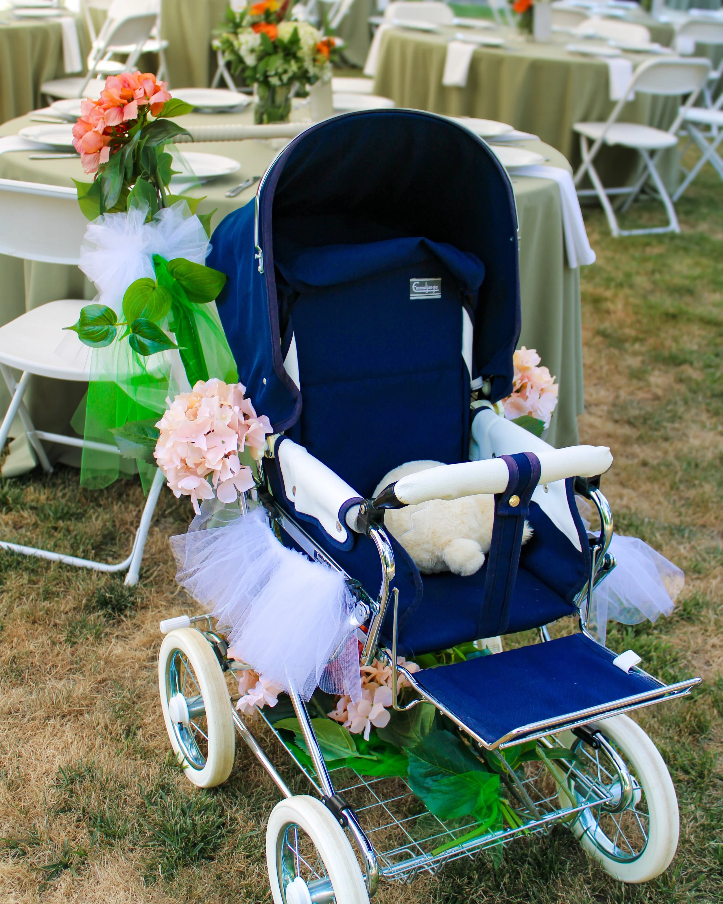 A blue baby stroller decorated with pink and peach flowers and green leaves, placed outdoors on a grassy area at a decorated event with round tables covered in beige tablecloths and white folding chairs in the background.