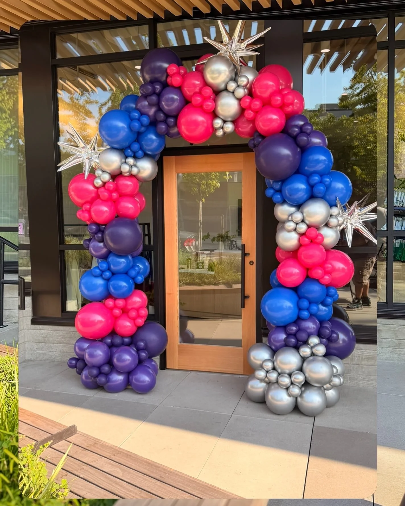 Balloon arch decorated with pink, purple, blue, and silver balloons, along with clear star-shaped balloons at the top corners, surrounding a wooden door entrance.