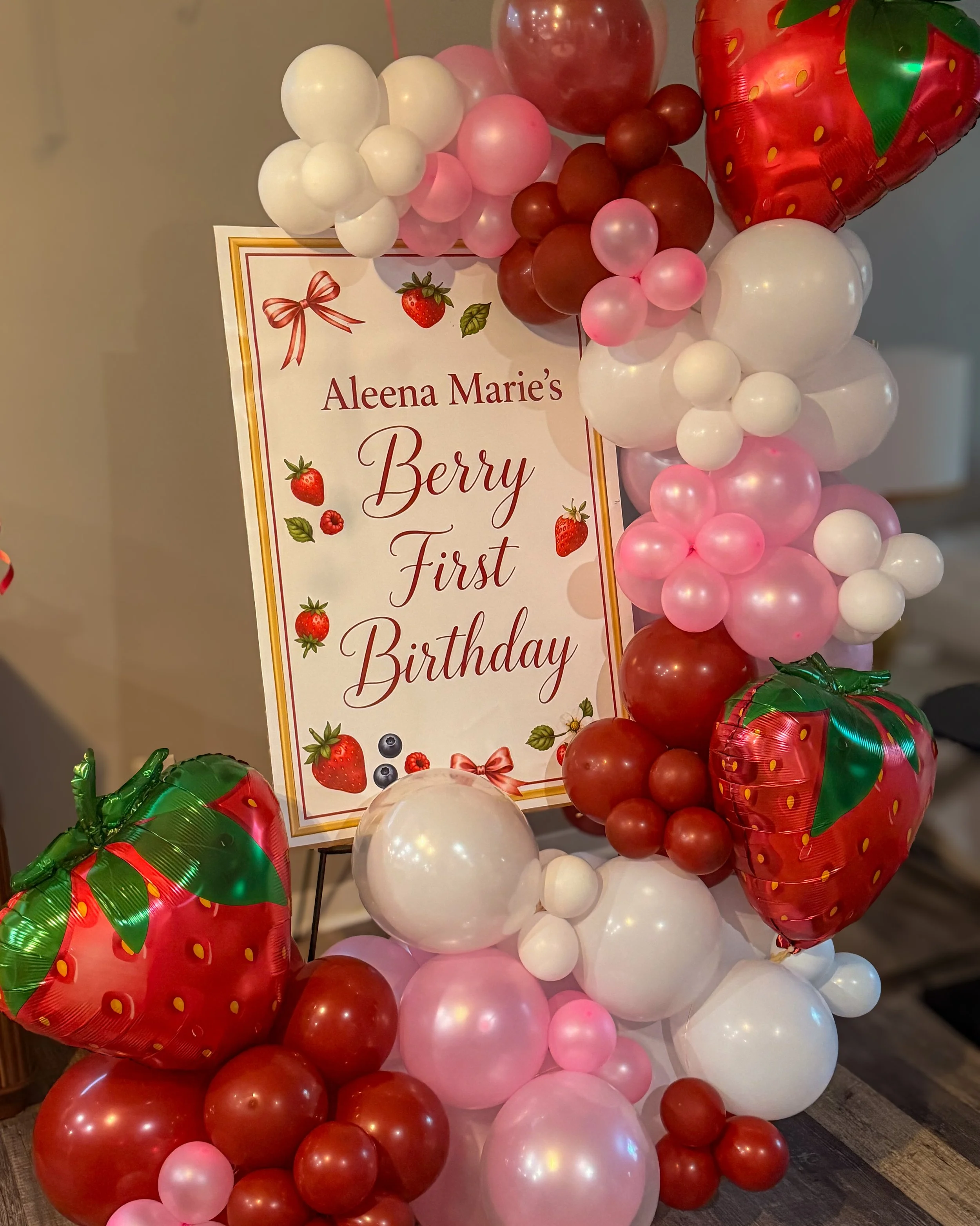 A colorful balloon decoration arrangement with a variety of pink, white, and red balloons, including strawberry-themed balloons, surrounding a sign that reads 'Aleena Marie's Berry First Birthday' with strawberry illustrations.
