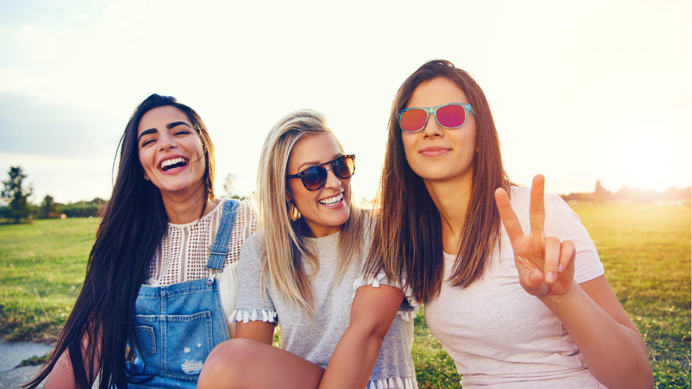 Trois jeunes femmes souriantes posent ensemble dans un champ, portant des lunettes de soleil, une d'elles fait un signe de paix avec les doigts.