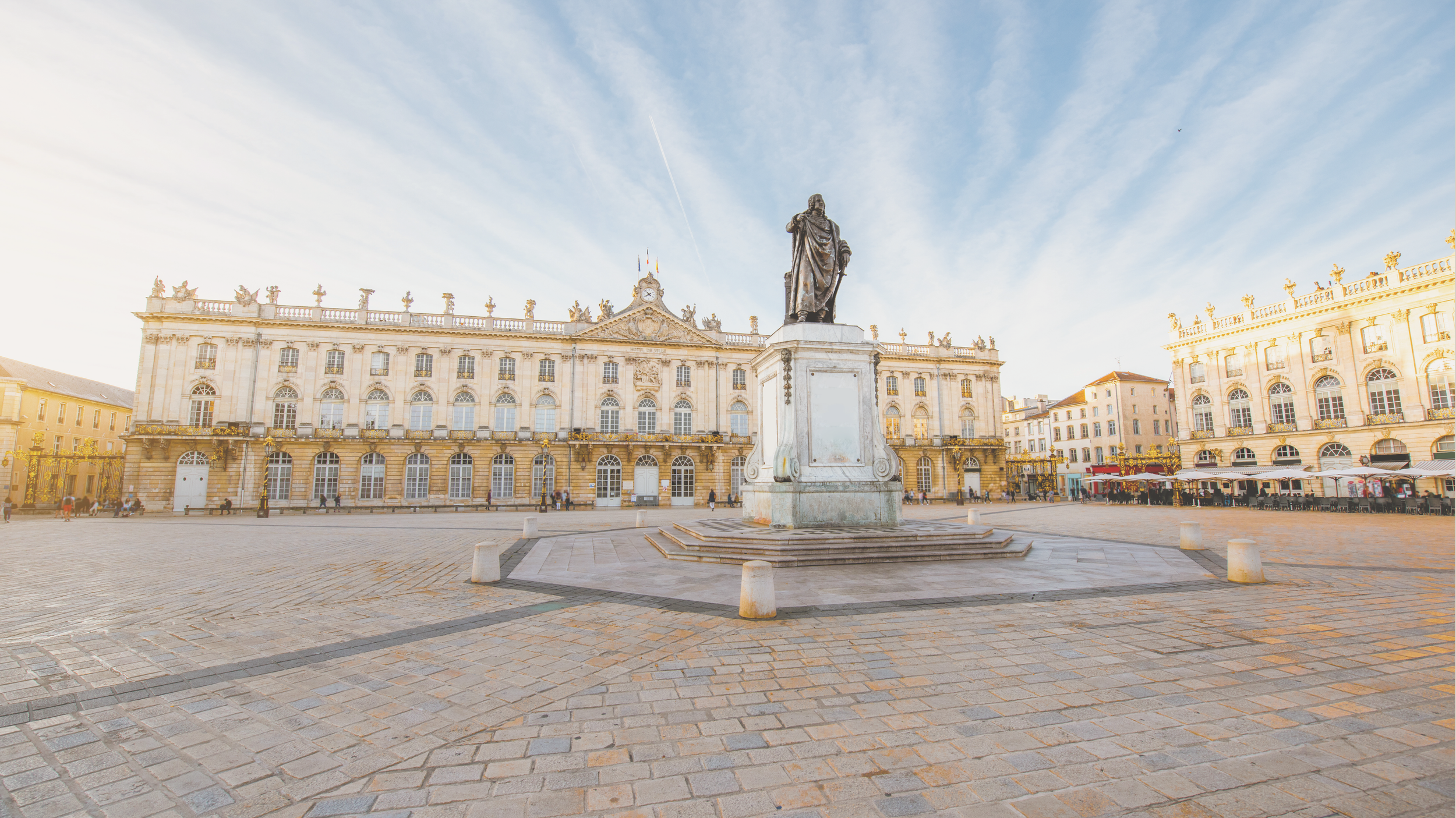 Place publique avec une statue au centre, bâtiment historique en arrière-plan, ciel dégagé, quelques passants et terrasses de cafés.
