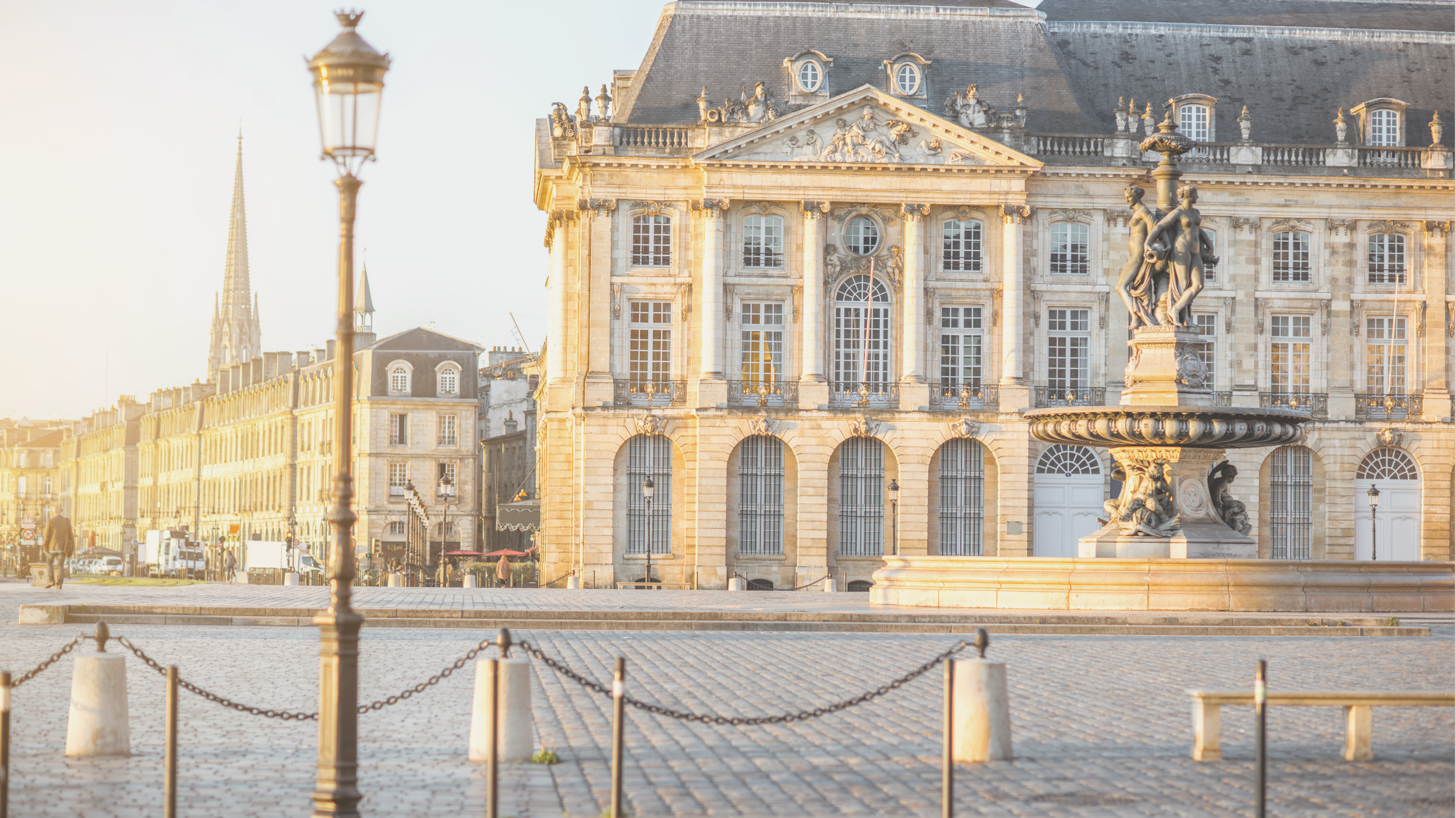 Bâtiment historique à l'architecture classique française avec fenêtres à petites croix, face à une fontaine ornée de sculptures de femmes et d'enfants, le tout éclairé par la lumière dorée du coucher de soleil.