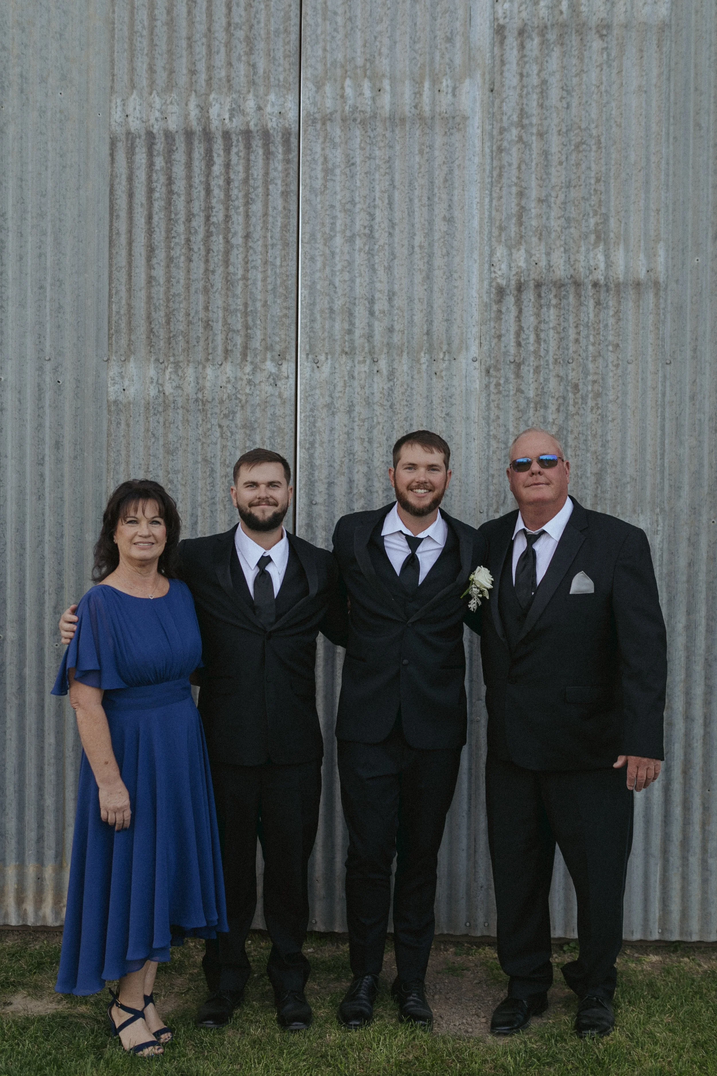 A group of four people standing outdoors in front of a metal wall. They are dressed in formal attire, with three men in black suits and ties, and a woman in a blue dress. The man on the far right is wearing sunglasses.