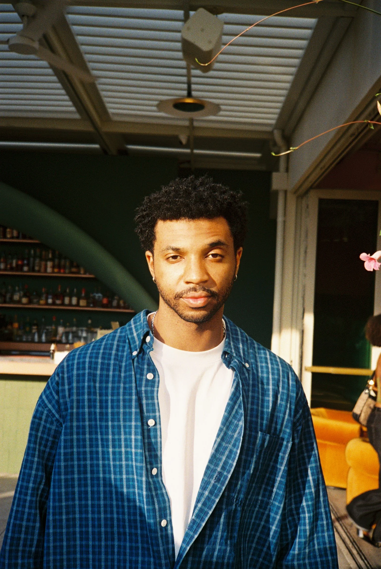 A young man with curly hair and earrings wearing a blue plaid shirt over a white t-shirt, standing indoors with a bar and green wall in the background.