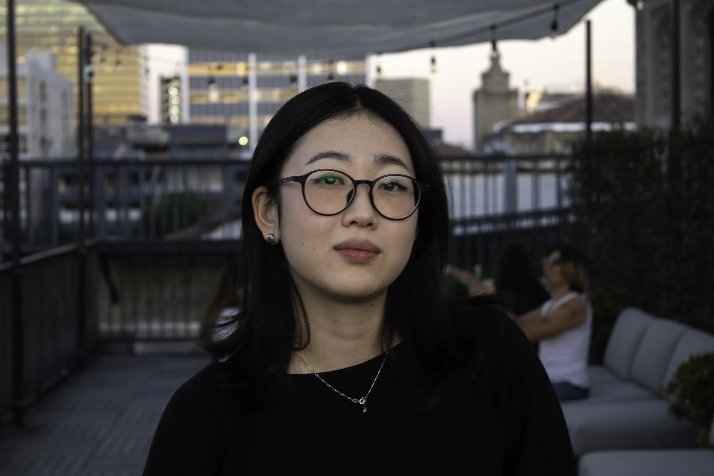 A woman with black hair, glasses, and a black top is sitting on a rooftop patio with a city skyline in the background at sunset.