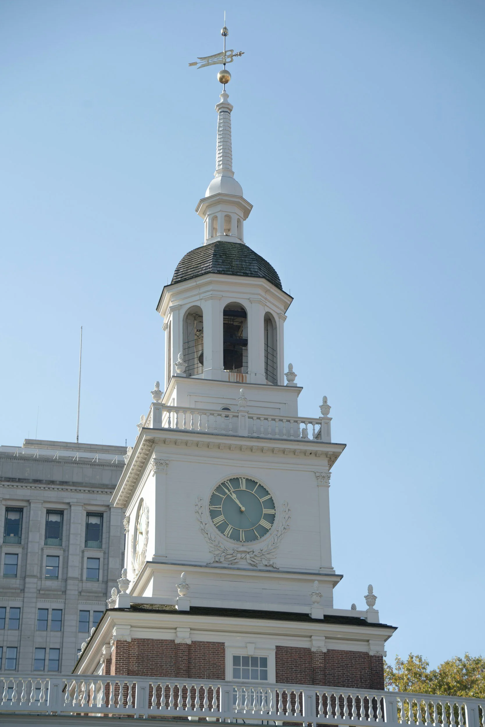A white clock tower with a black roof, decorative details, and a weather vane at the top, set against a clear blue sky.