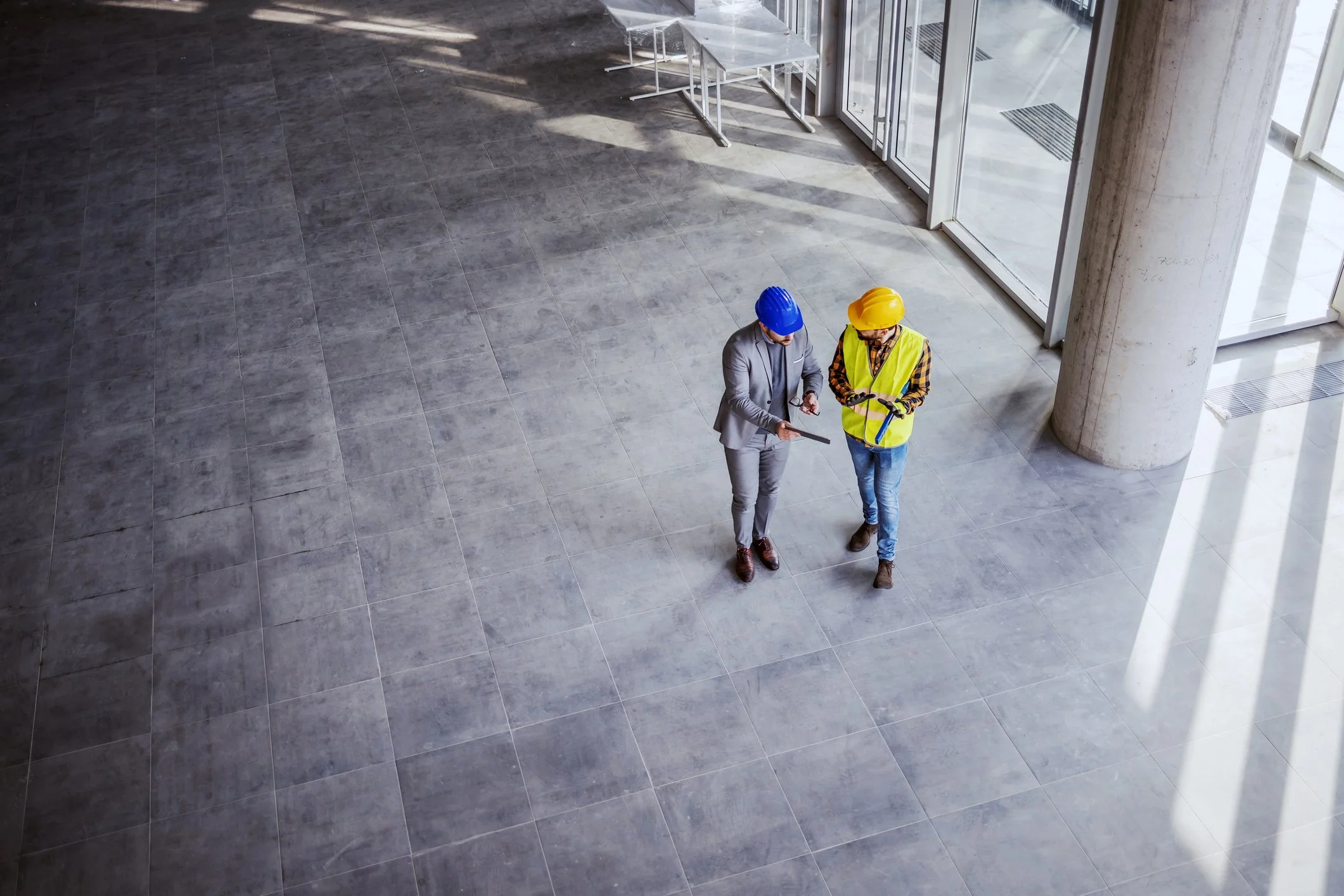 Two men wearing safety helmets and one wearing a high-visibility vest are standing and talking inside a modern building with large glass windows and a grey tiled floor.