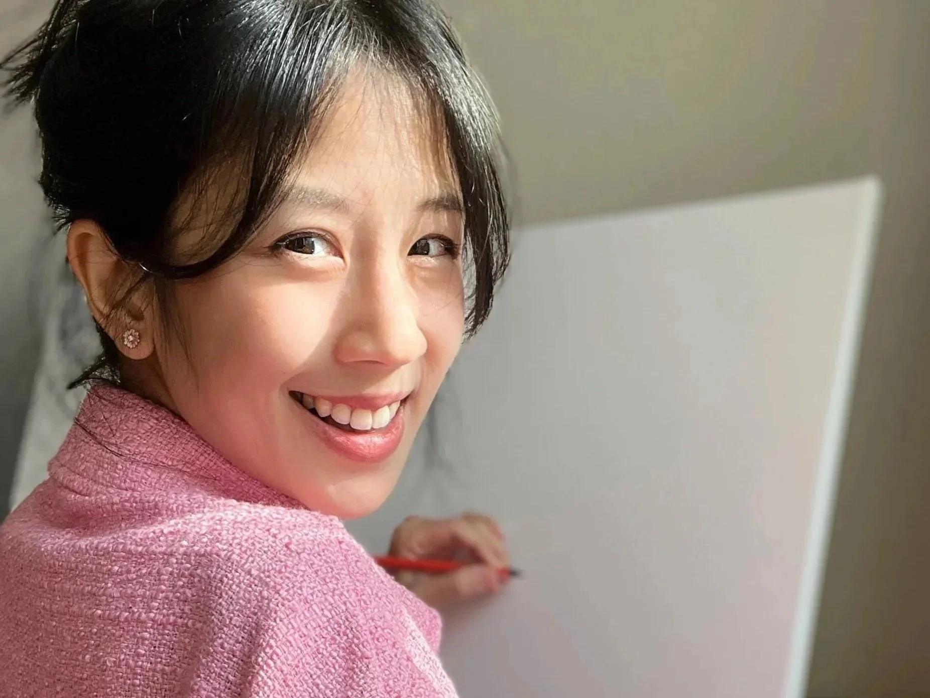 A vietnamesse artist with black hair  smiles while looking at the camera, wearing a pink textured jacket, with a whiteboard or easel in the background. She is sketching on the canvas.