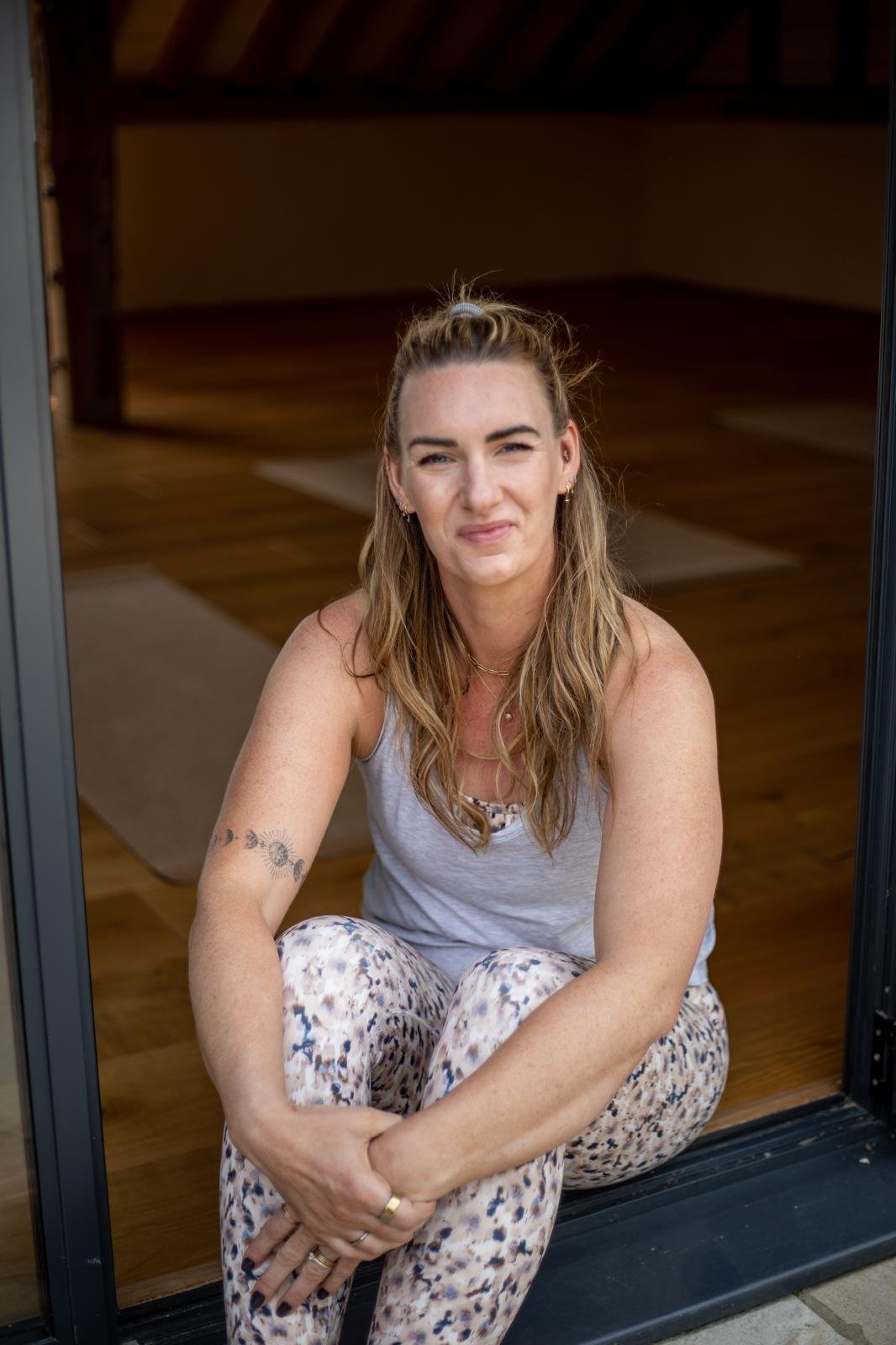 A woman sitting at the doorway of a wooden house, squatting on the ground, looking at the camera with a slight smile. She has long wavy hair, tattoos on her arm, and is wearing a sleeveless top and patterned pants.