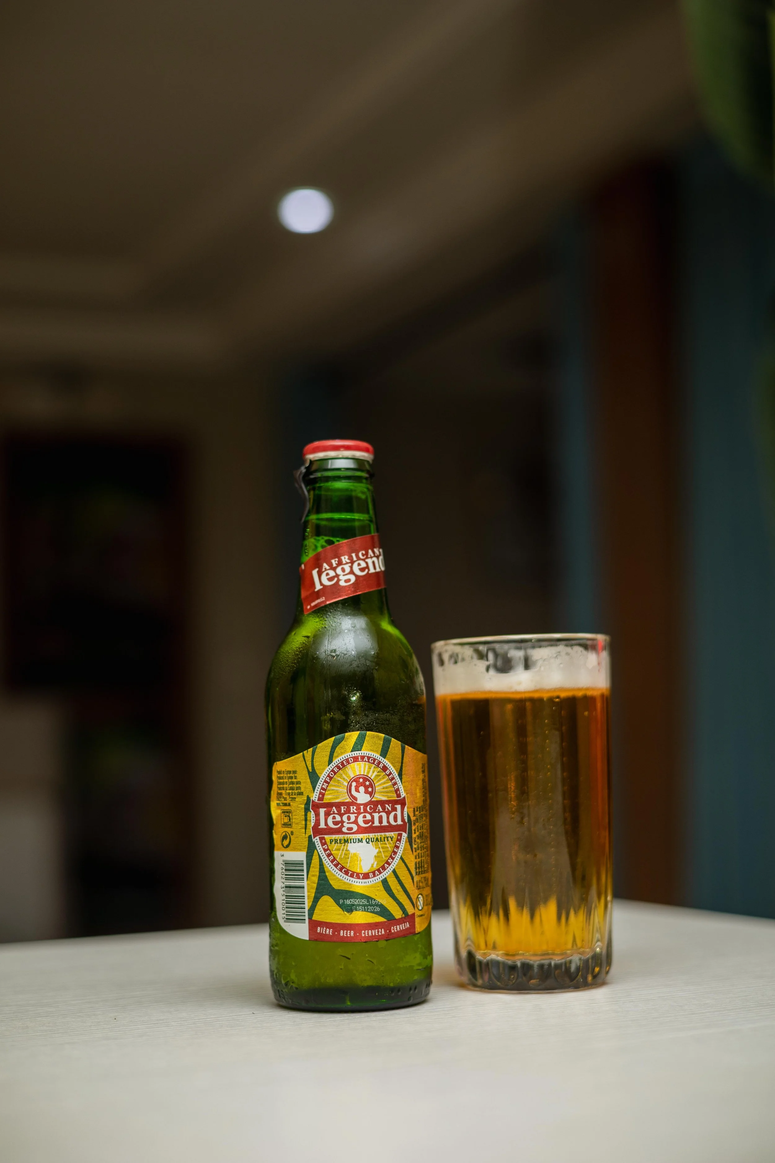 A green bottle of African Legend beer with a red and yellow label next to a tall glass of beer on a white surface, with a blurred background.