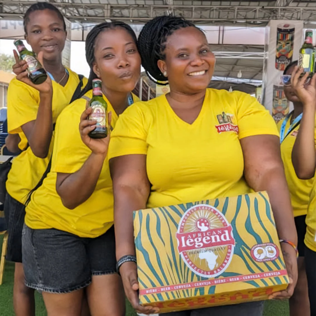 Group of women in yellow shirts standing together at an event, smiling and holding beer bottles, with one woman holding a colorful box labeled 'African Legend'.