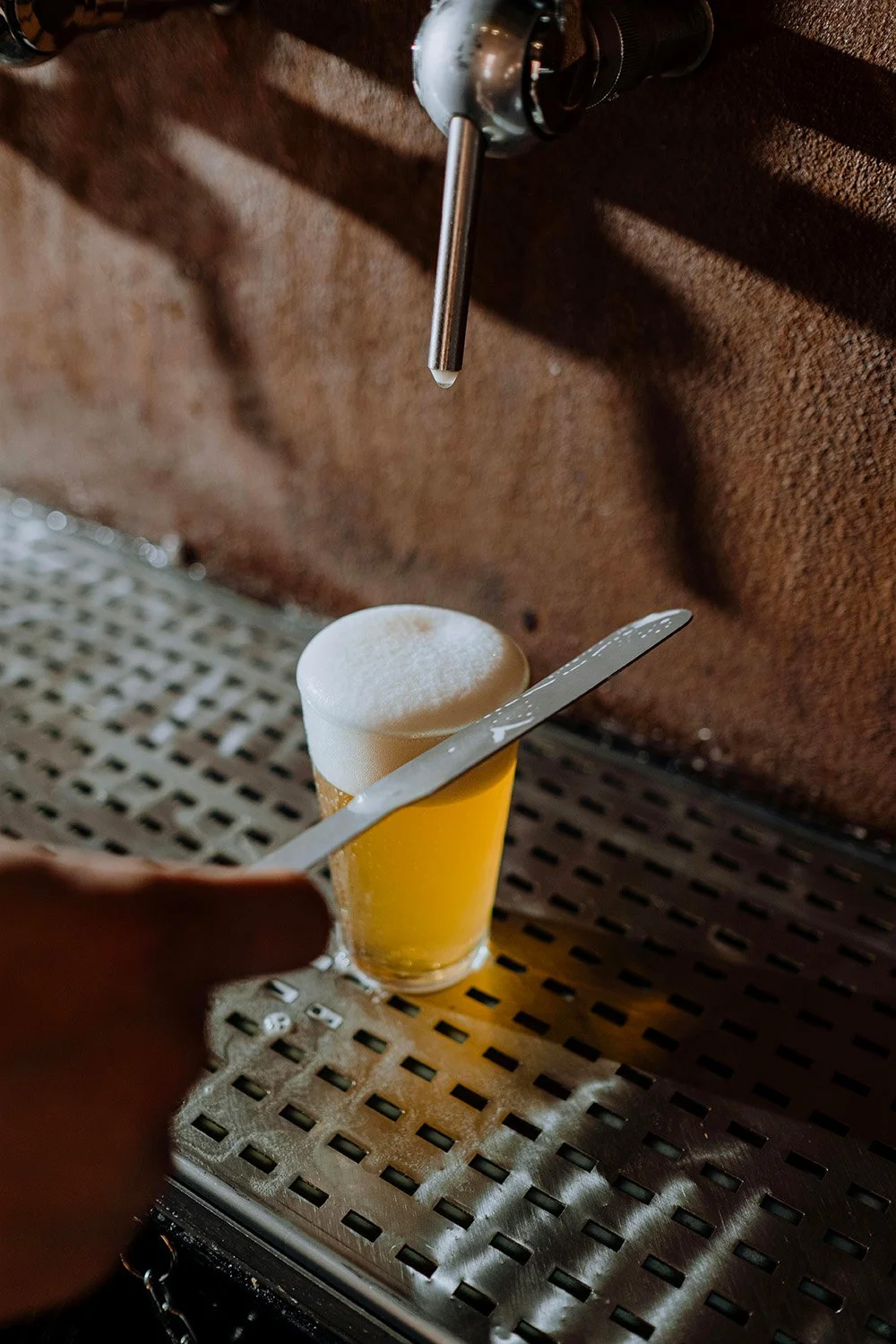 A person holds a flat metal spatula and a tool with foam on top, positioned above a pint of pale, cloudy beer with foam on top, under a tap pouring beer, on a metallic tray.