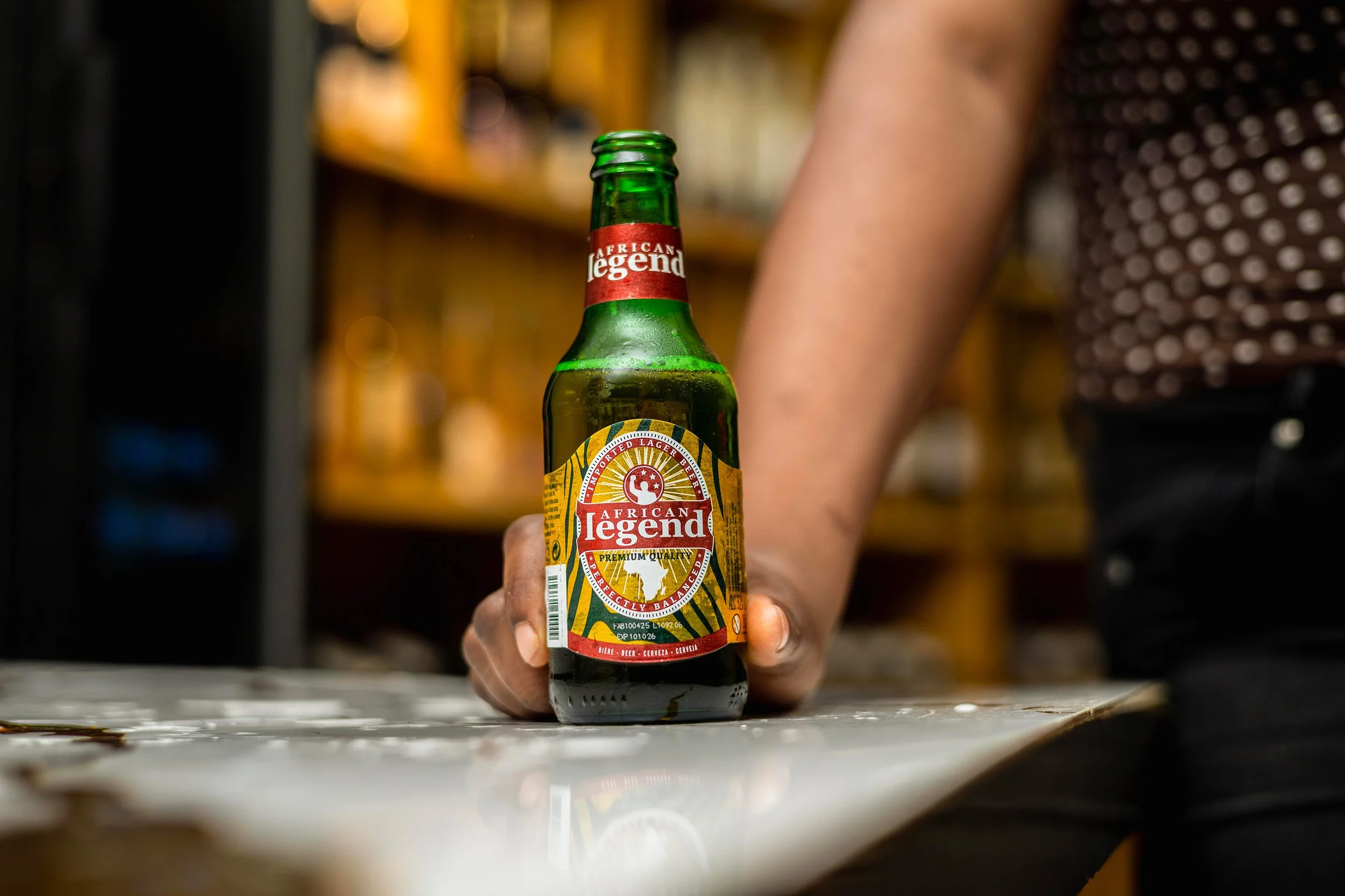 A hand holding a green glass bottle of African Legend beer on a table, with a blurred background of shelves and a person wearing a brown and white polka dot shirt.