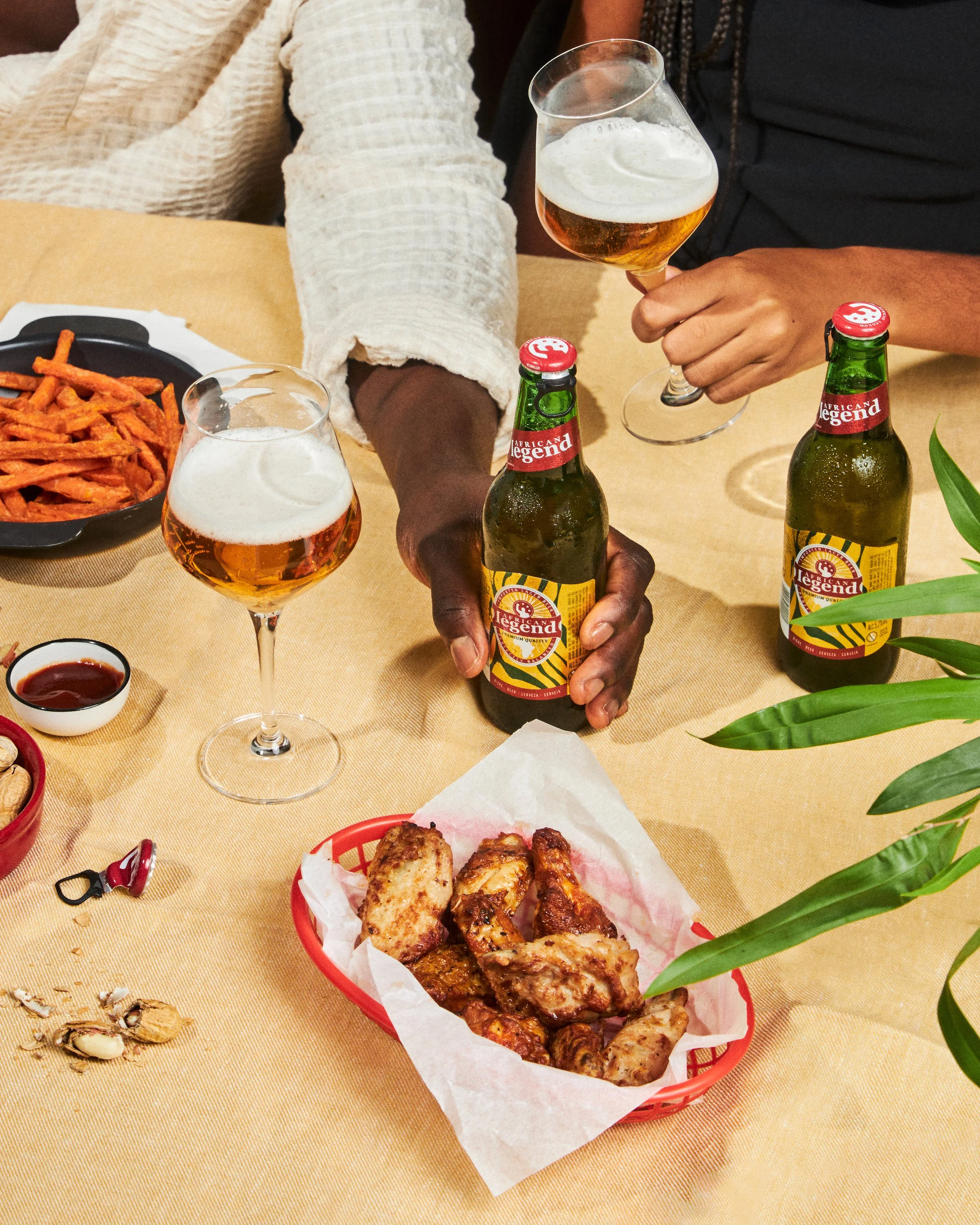 A table with food and drinks at a casual gathering. Two hands holding glasses of beer, one person with dark skin and another wearing a black shirt. On the table, there is a red basket of fried chicken, a small bowl of dipping sauce, a black plate of sweet potato fries, bottled beer with a Tiki Torch cap, and some scattered peanuts and shells. The background includes a plant with green leaves.