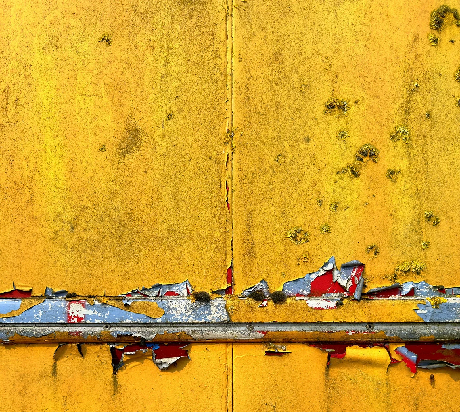 Close-up of a weathered, yellow metal surface with peeling paint, rust spots, and some underlying red, white, and gray layers showing through