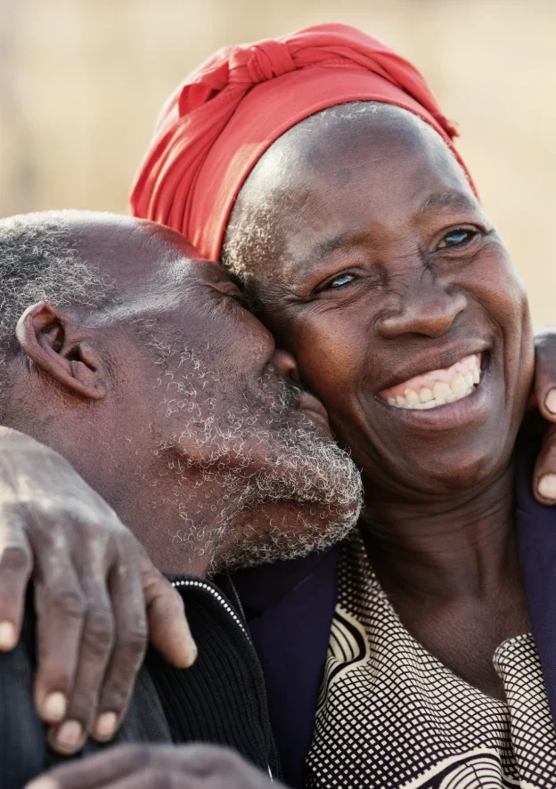 An elderly man with gray hair and beard embracing a smiling woman wearing a red headscarf.