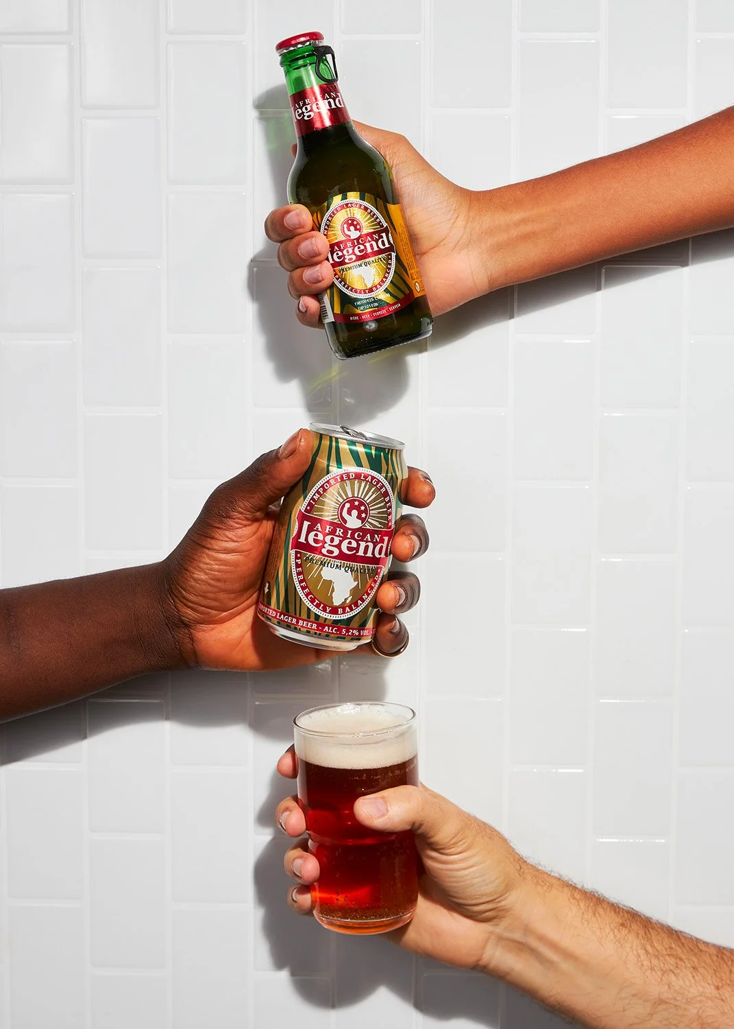 Three hands holding different alcoholic beverages: a green bottle of African Legend beer, a can of African Legend beer, and a glass of beer, against a white tiled wall.