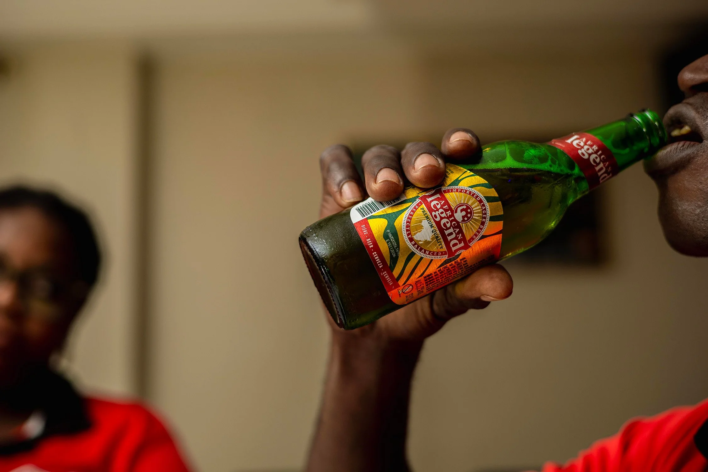 A person with dark skin holding a green glass bottle of Bigelow brand beer to their mouth, about to drink, with a blurred background.