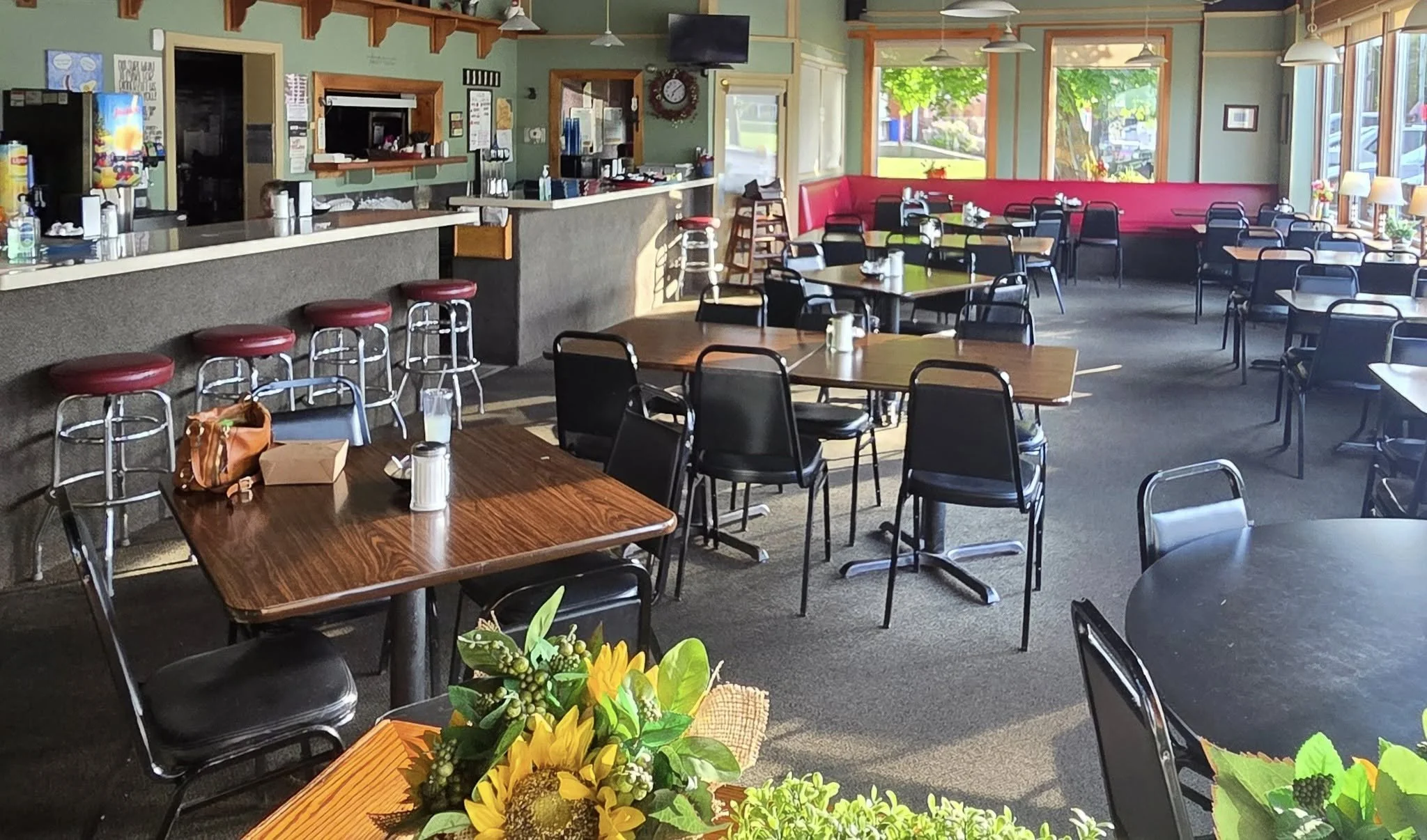 Empty restaurant with wooden tables and black chairs, sunlight streaming through large windows, flowers on some tables, a red cushioned bench along the wall, and a bar area in the background.
