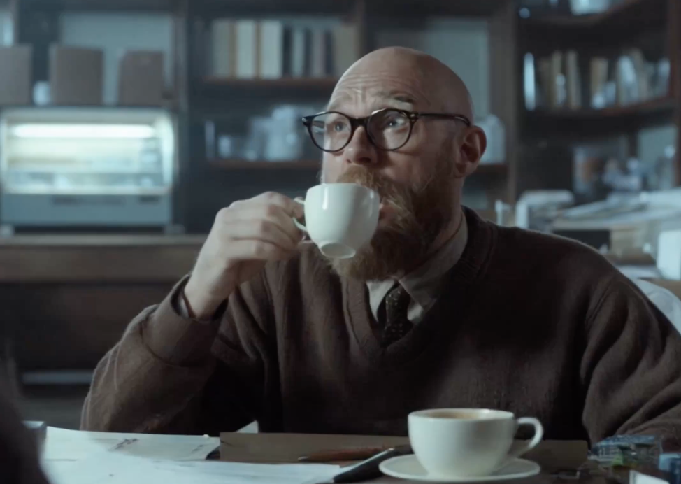 A man with glasses and a beard drinking coffee in a cafe or library setting.