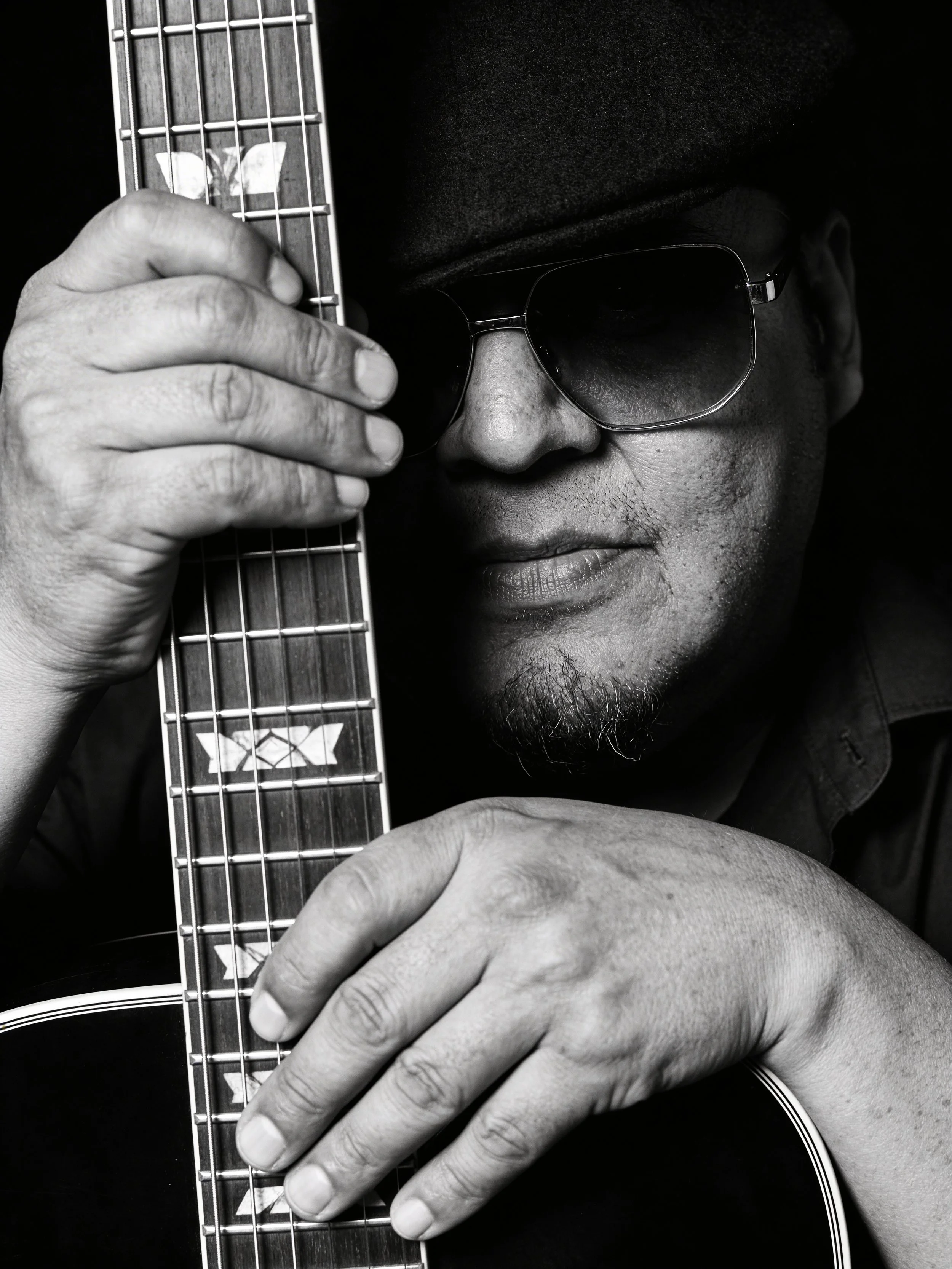 A man wearing sunglasses and a cap, holding an acoustic guitar close to his face, with one hand on the fretboard and the other on the body of the guitar, in black and white.