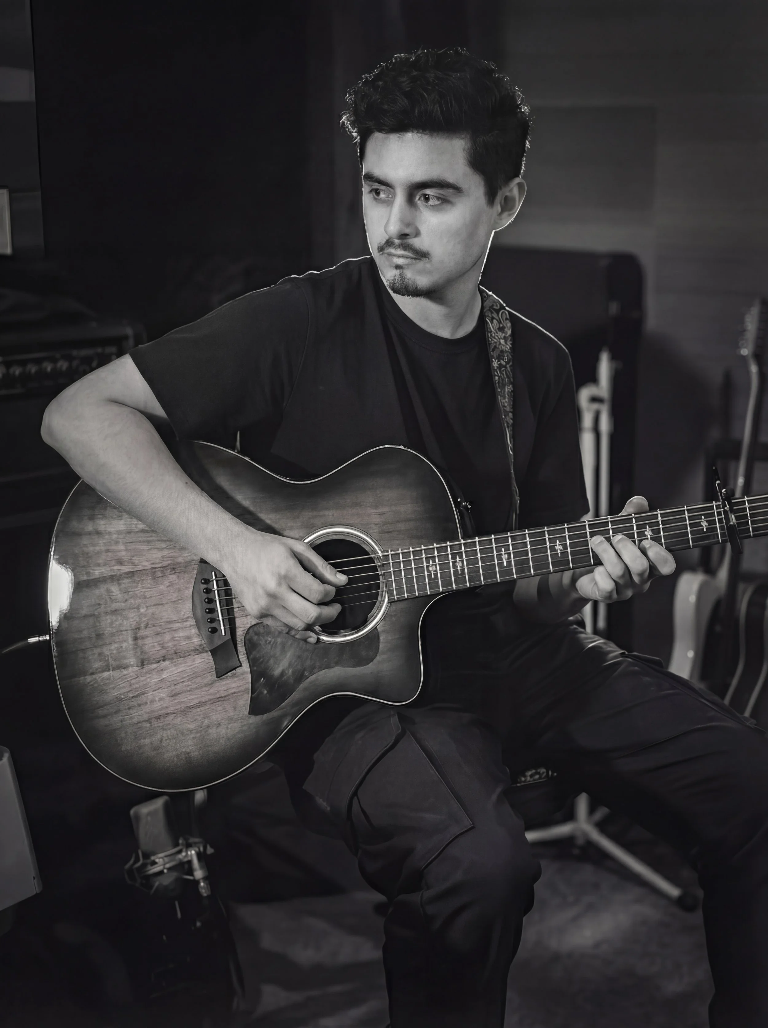A young man with dark hair and slight facial hair, wearing a black t-shirt and pants, is playing an acoustic guitar in a studio setting.