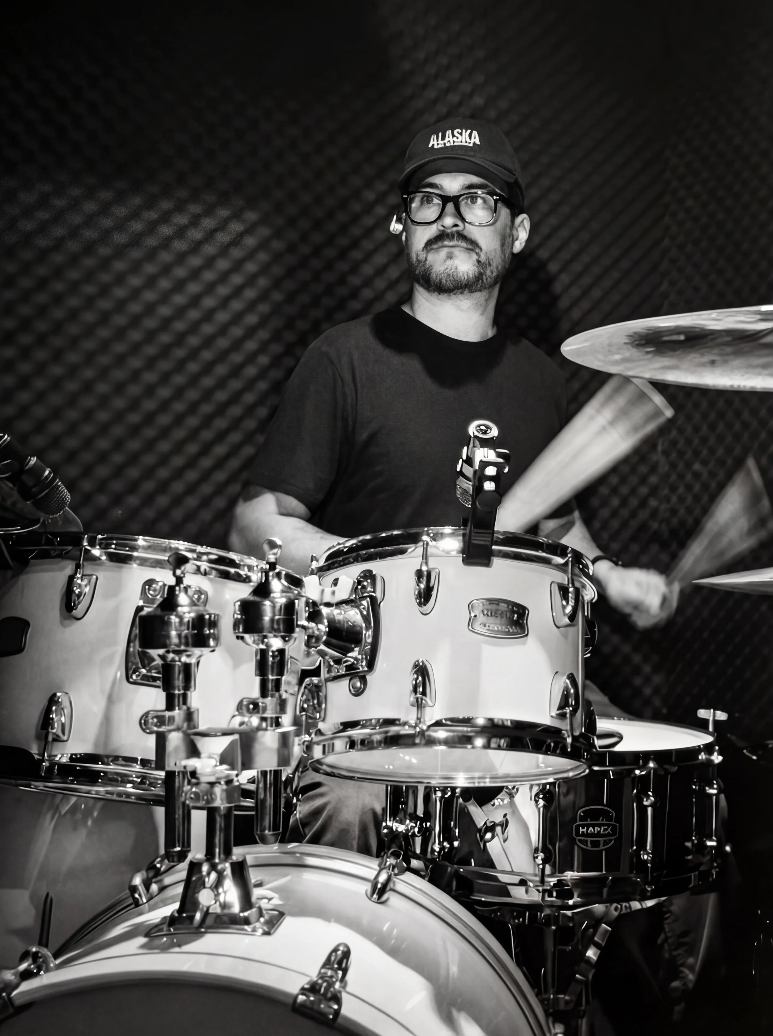 A man with glasses and a beard, wearing a cap and t-shirt, playing a drum set in a music studio.